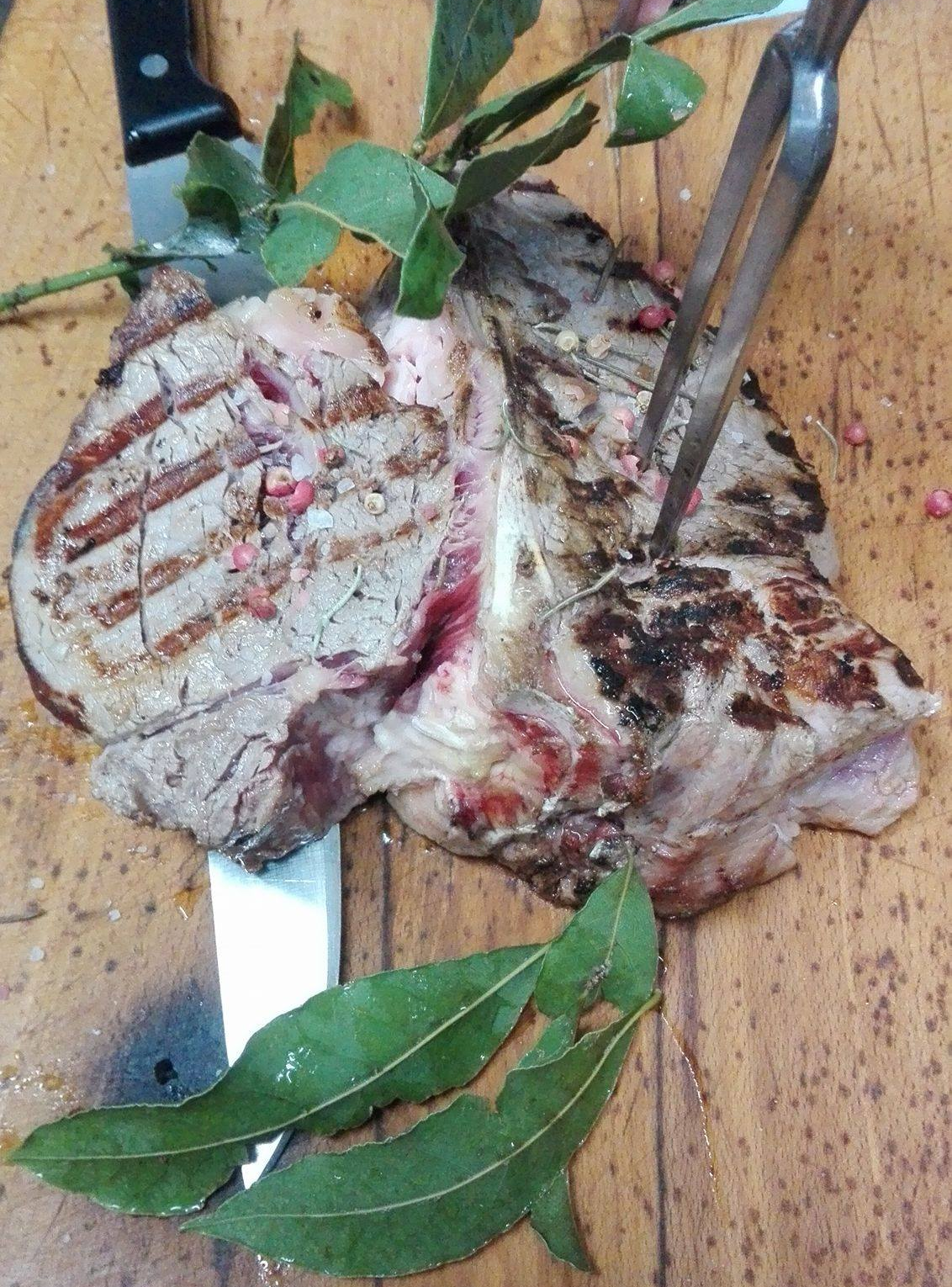Bistecca grigliata su una superficie di legno con coltello e pinze, guarnita con foglie di alloro.