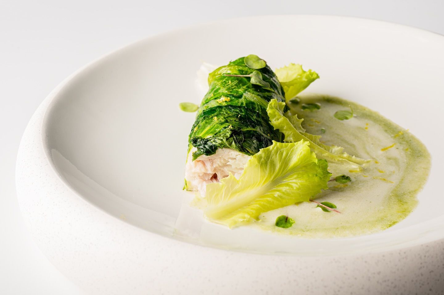 Piatto bianco con una foglia di cavolo arrotolata ripiena di pesce, circondata da salsa e lattuga.
