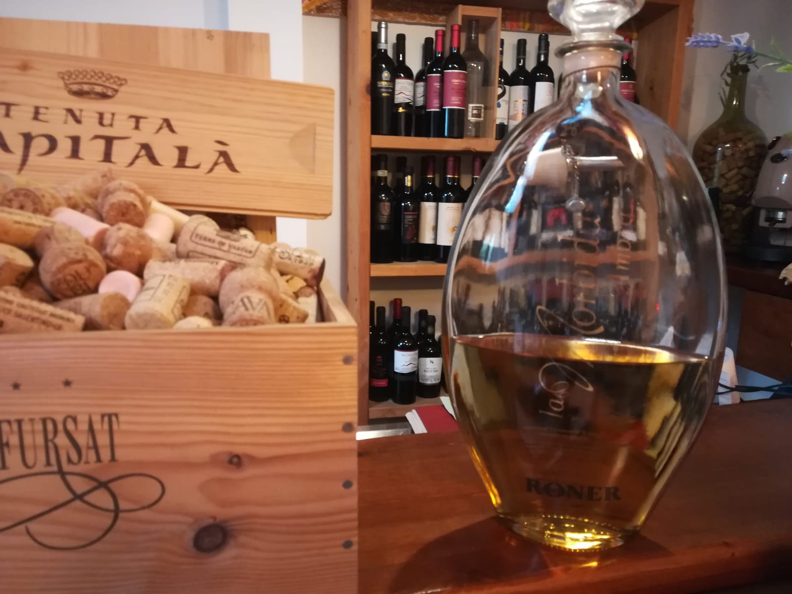 Una scatola di legno piena di tappi di sughero accanto a una caraffa di liquido color ambra, e sullo sfondo delle bottiglie su uno scaffale.