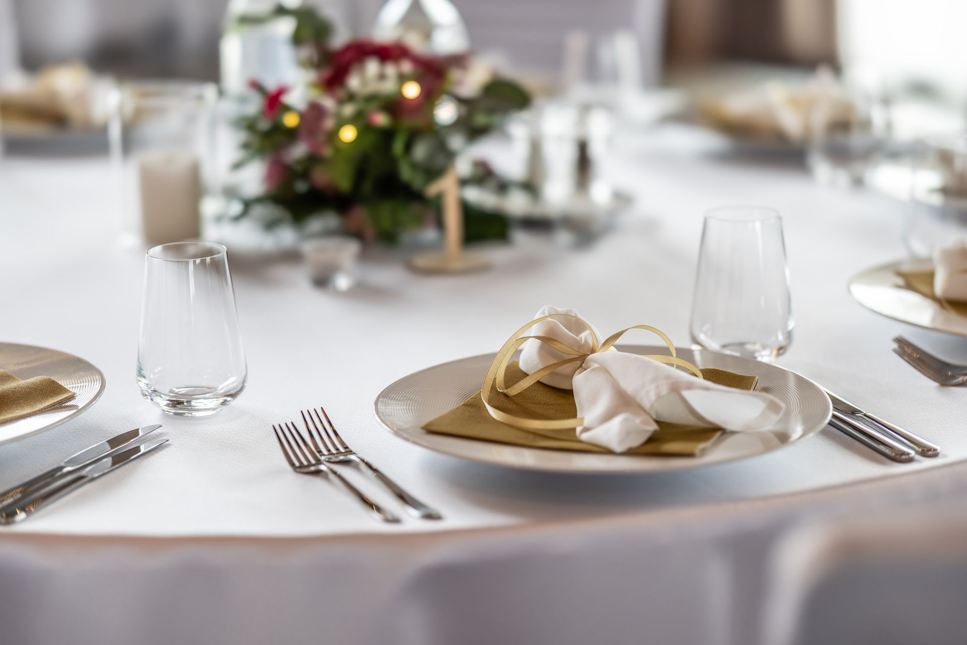 Tavolo bianco apparecchiato per un evento formale con dettagli dorati, piatti, posate e centrotavola floreale.