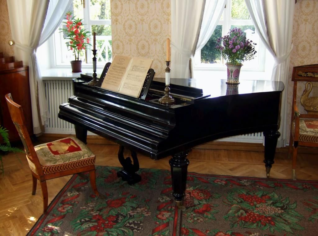 A black grand piano with a music book and candle sits on a floral rug in a room with white curtains and two windows.