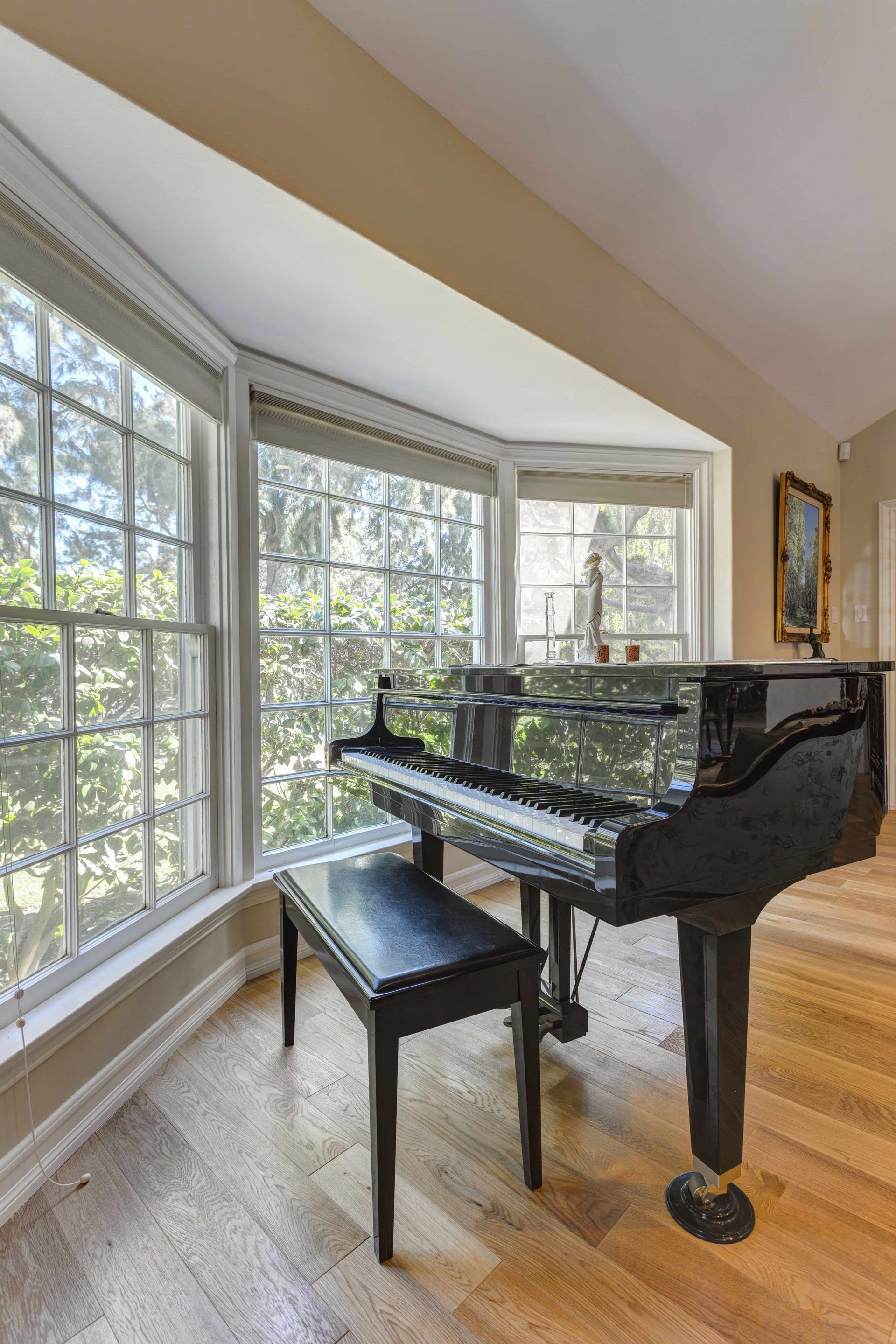 A black grand piano with a matching bench sits on a wooden floor in a sunlit bay window area with white trim.