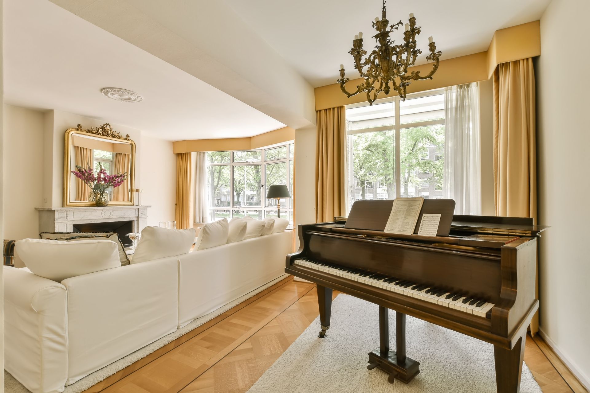 A living room with a grand piano, a large white sofa, a fireplace with a mirror above it, and tall beige curtains.