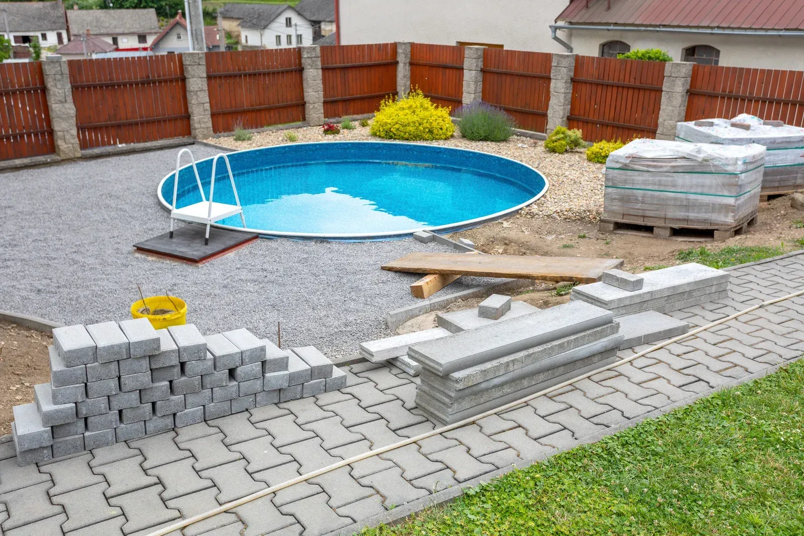 Backyard with pool, gravel, paving stones, wooden fence, and construction materials.