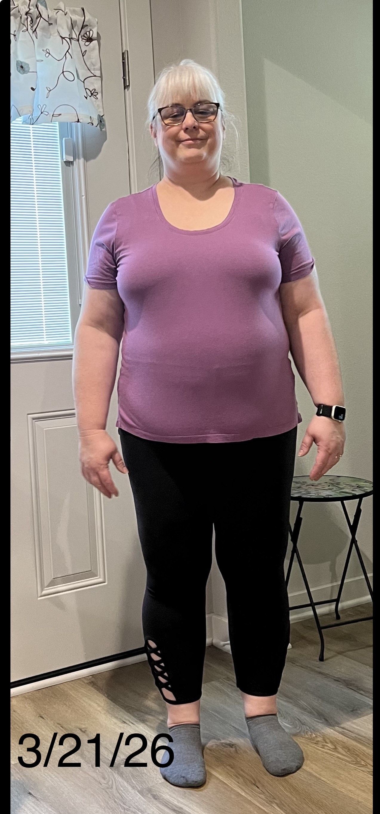 Woman standing indoors, wearing purple shirt and black capris, smiling.