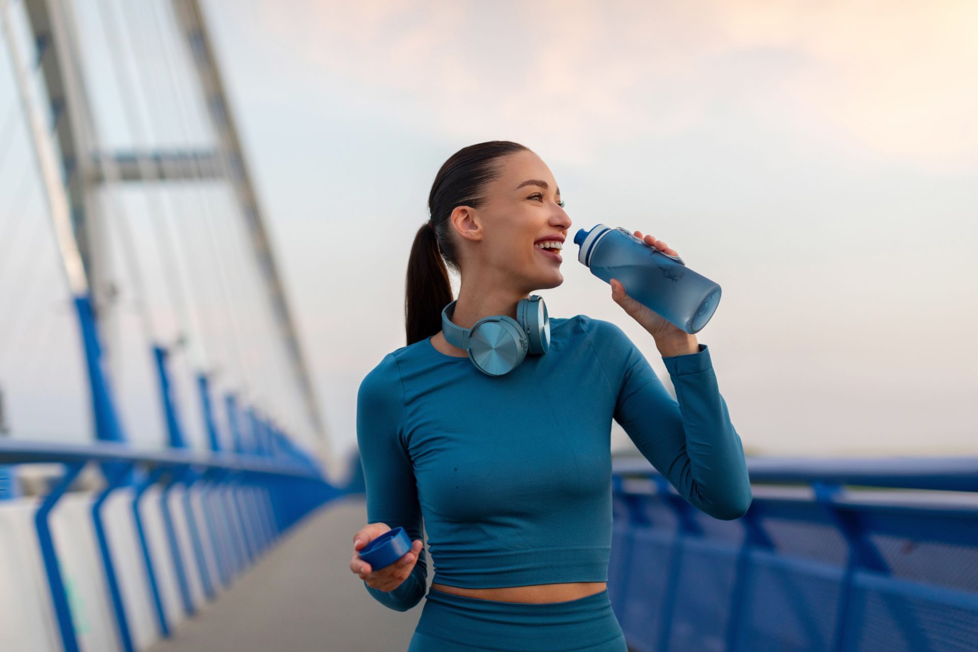 Woman in teal workout gear drinking water on a bridge while holding a phone, with headphones around her neck