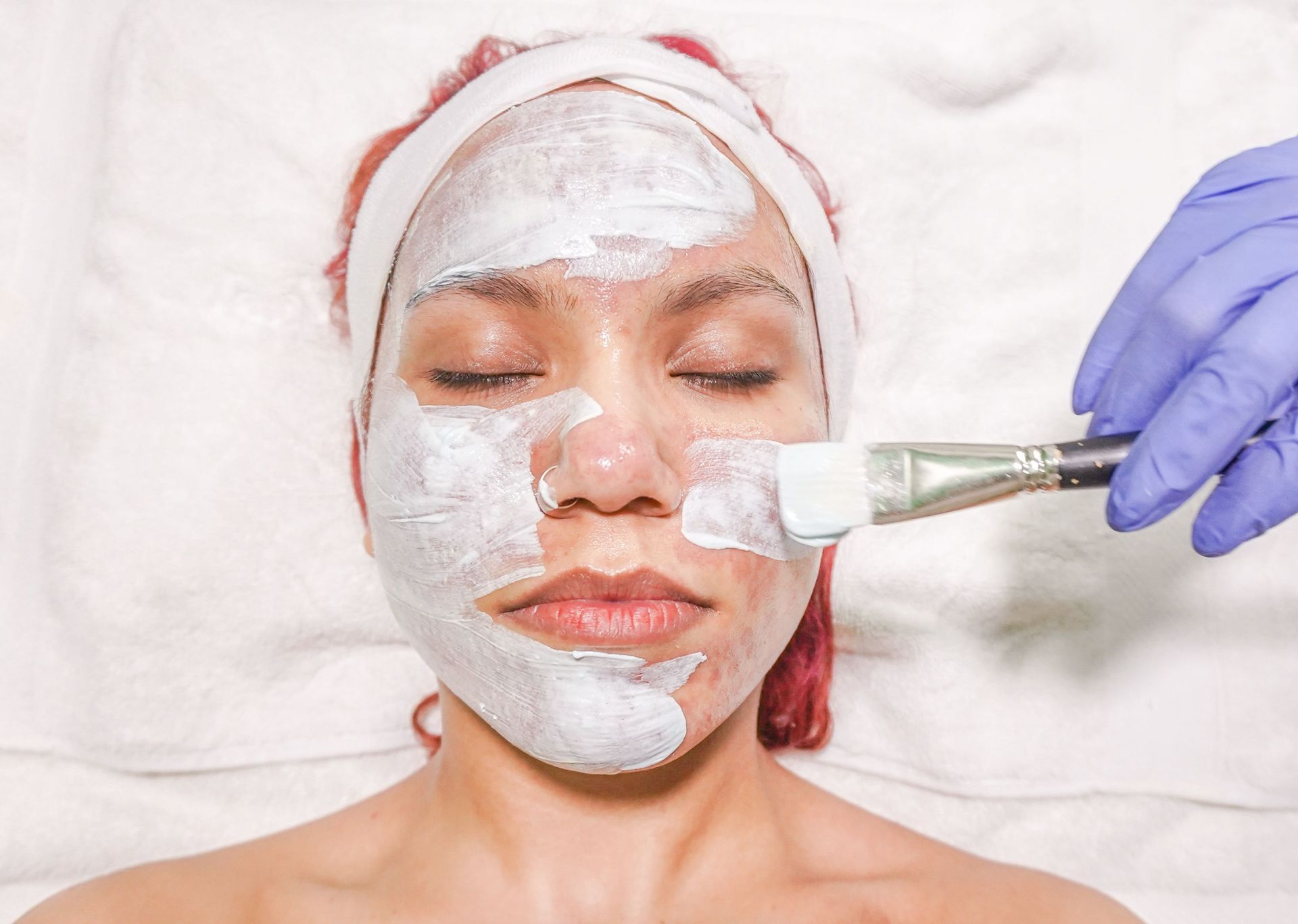 Woman receiving a white facial mask treatment with a brush while lying on a spa bed