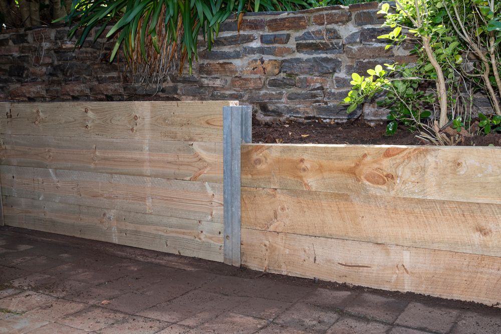 Wooden retaining wall with metal posts beside a paved patio and stone garden bed. — KLS Fencing In Normanton, QLD