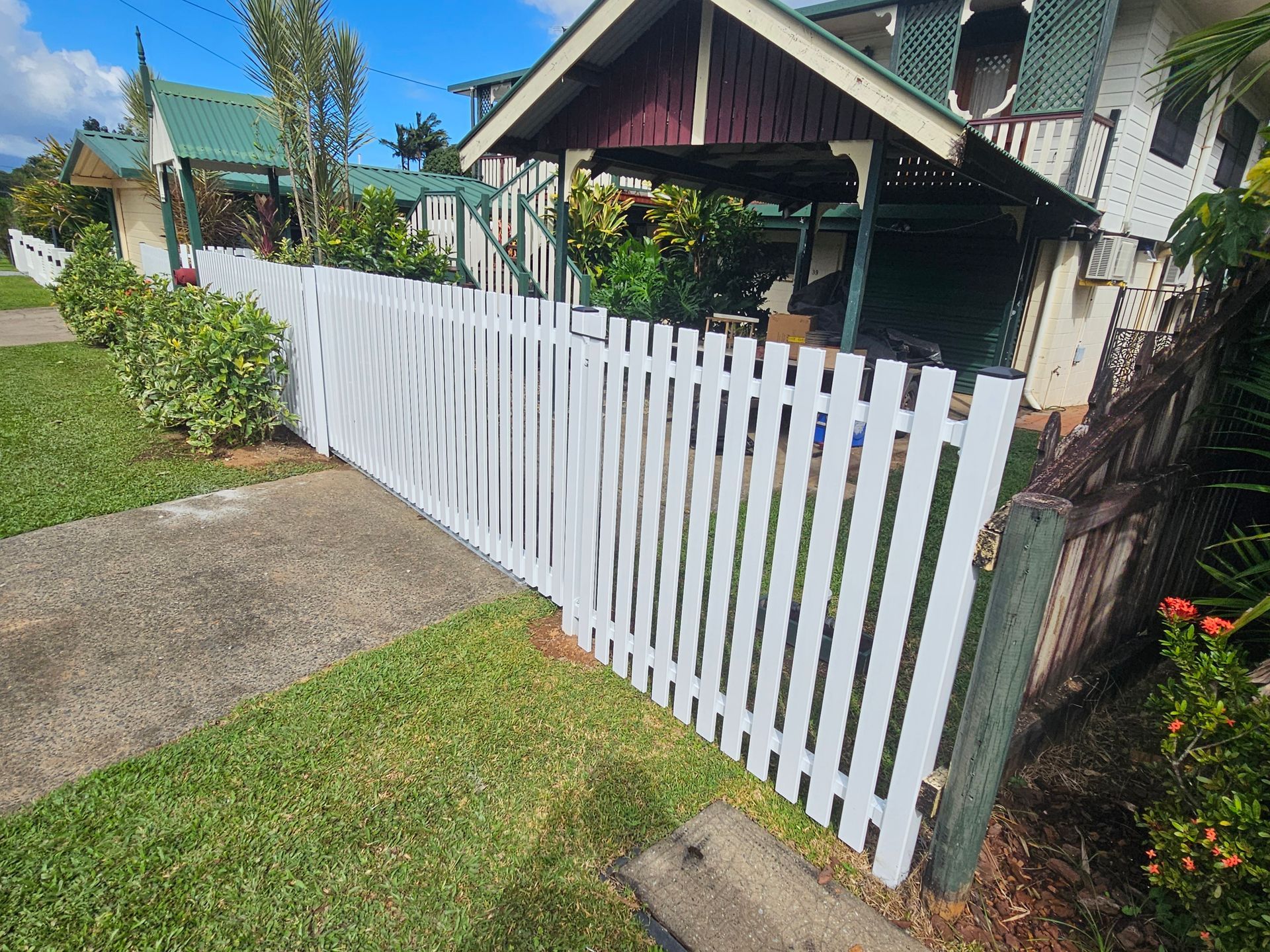 White picket fence along a path beside a small house and garden on a sunny day