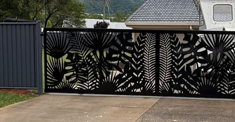 Black metal driveway gate with large tropical leaf cutouts beside a fenced driveway outdoors — KLS Fencing In Mossman, QLD