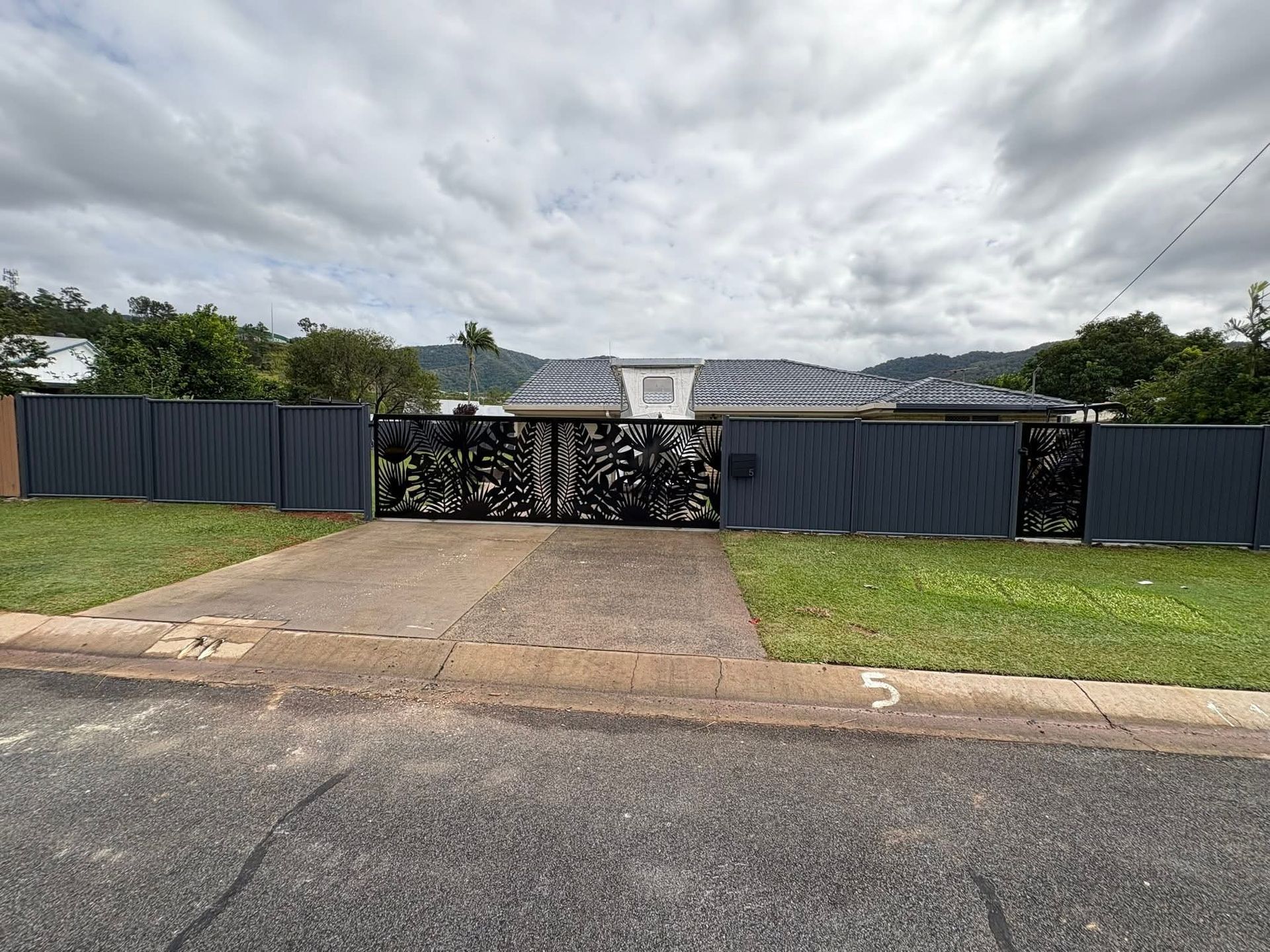 Gated driveway with black fence and metal gate in front of a house, under a cloudy sky.