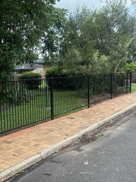 Black metal fence beside a brick sidewalk, with trees and a house behind it on a quiet street.
