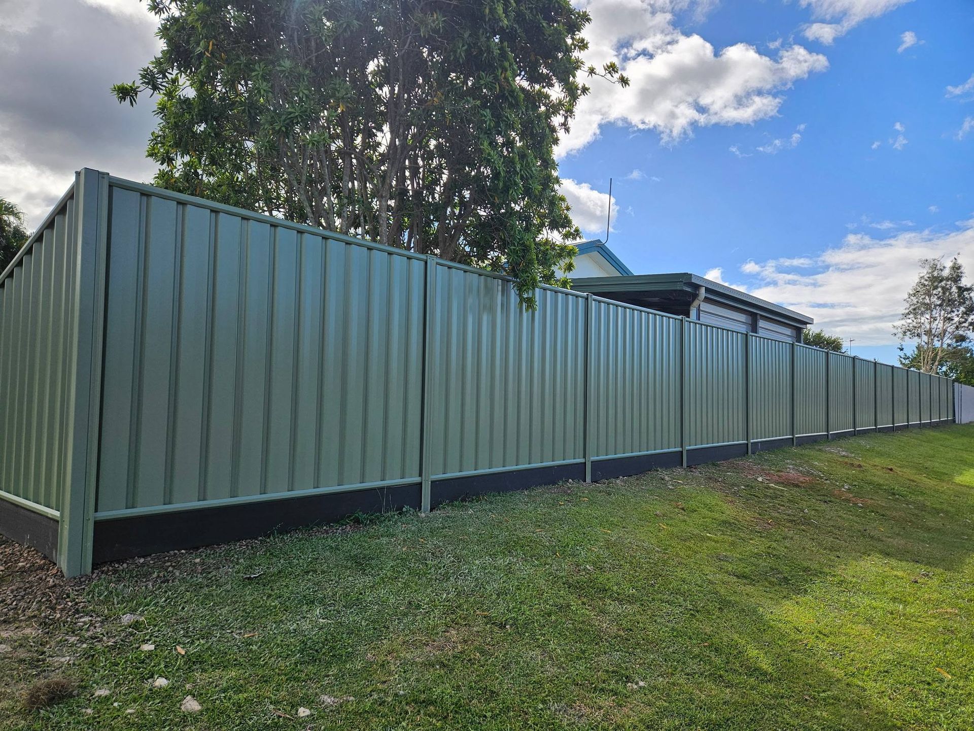 Long corrugated metal fence along a grassy yard under a blue sky with clouds. — KLS Fencing In Edmonton, QLD