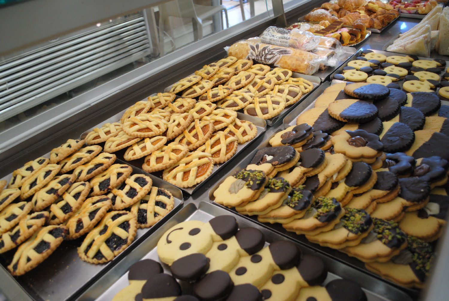 Biscotti assortiti decorati, esposti in una vetrina da pasticceria, con disegni a griglia, a girasole e ricoperti di cioccolato.