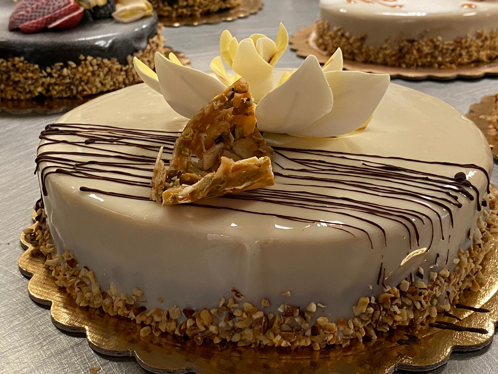 Torta rotonda ricoperta di crema, con glassa al cioccolato, bordi ricoperti di noci e guarnizione di cioccolato bianco, servita su un vassoio.
