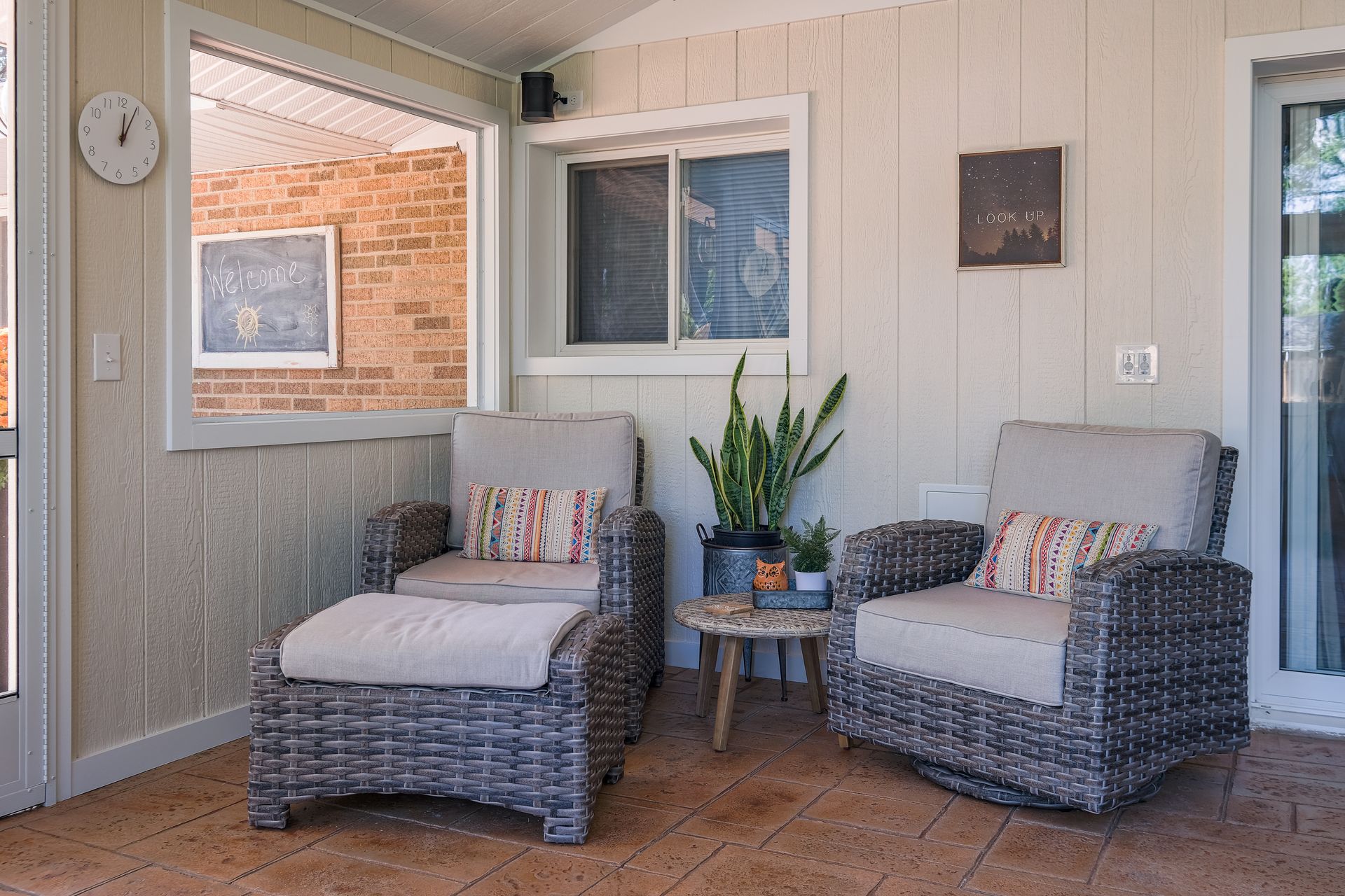 Two wicker chairs with a footstool on a covered patio; a small table and a plant sit nearby.