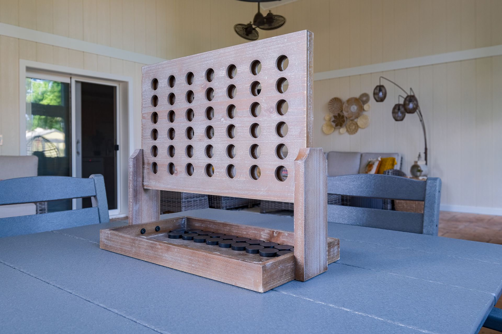 Giant Connect Four game on a gray table outdoors, with a neutral-toned backdrop of a porch and trees.
