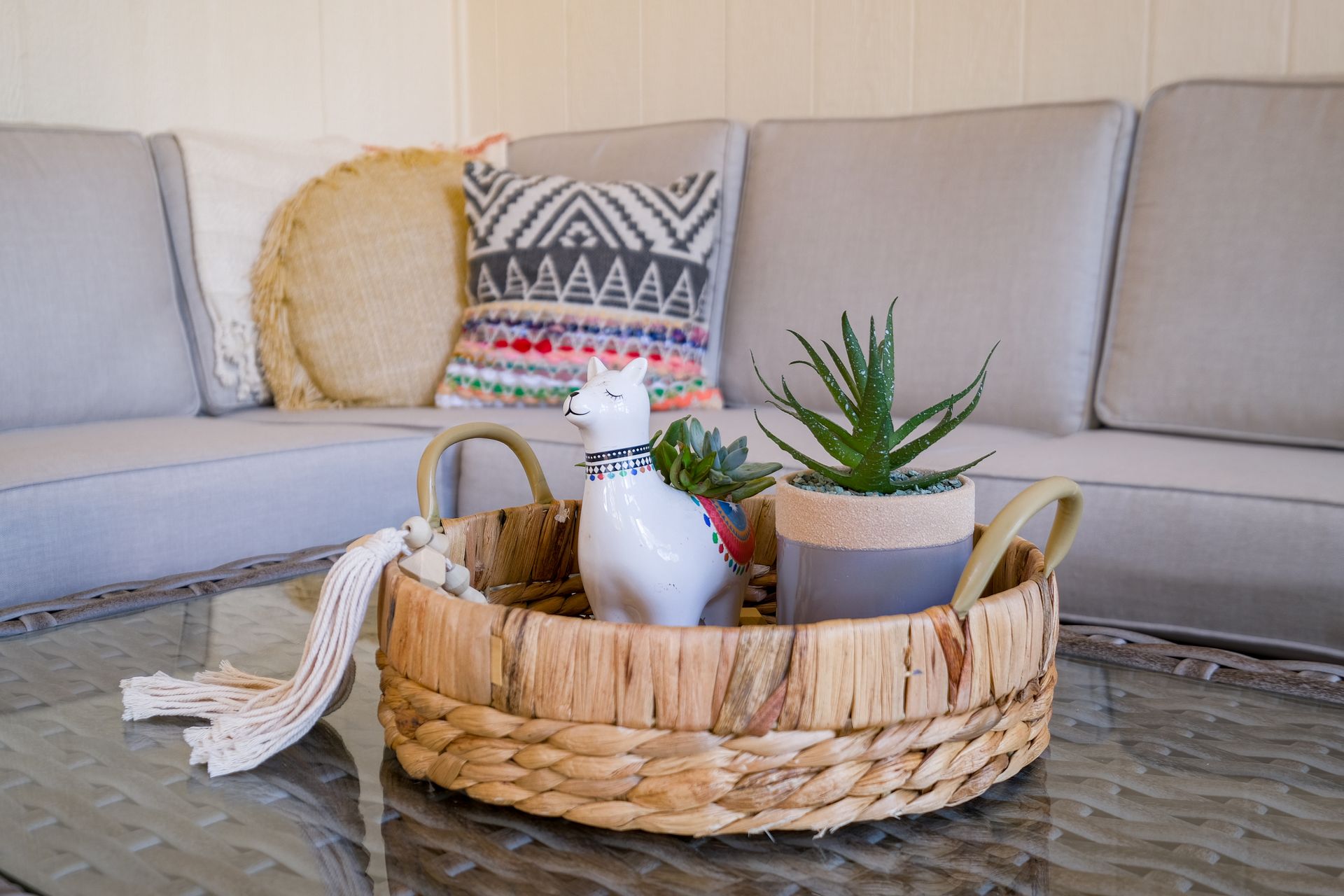 Wicker tray with a white llama figurine and succulent on a glass table, gray sofa in the background.