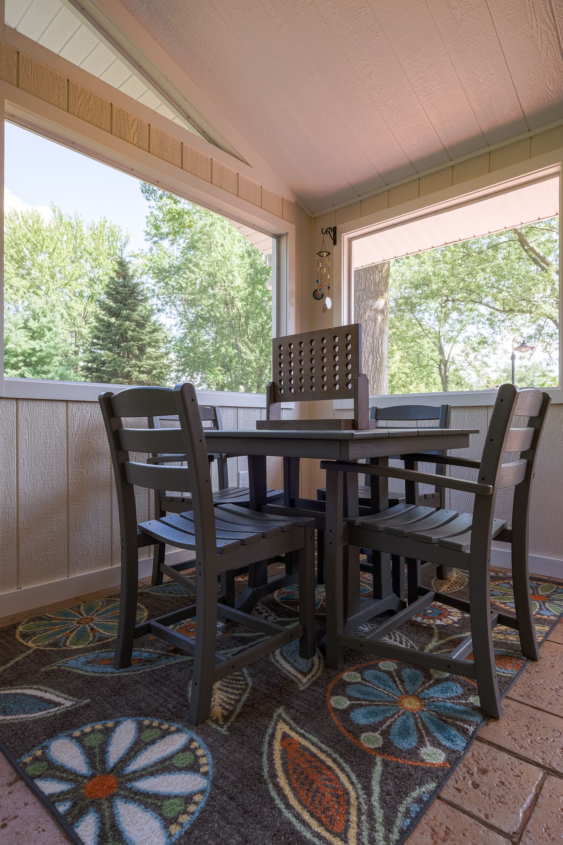 A screened-in porch with a gray square table and chairs on a floral rug, with a view of trees through the windows.
