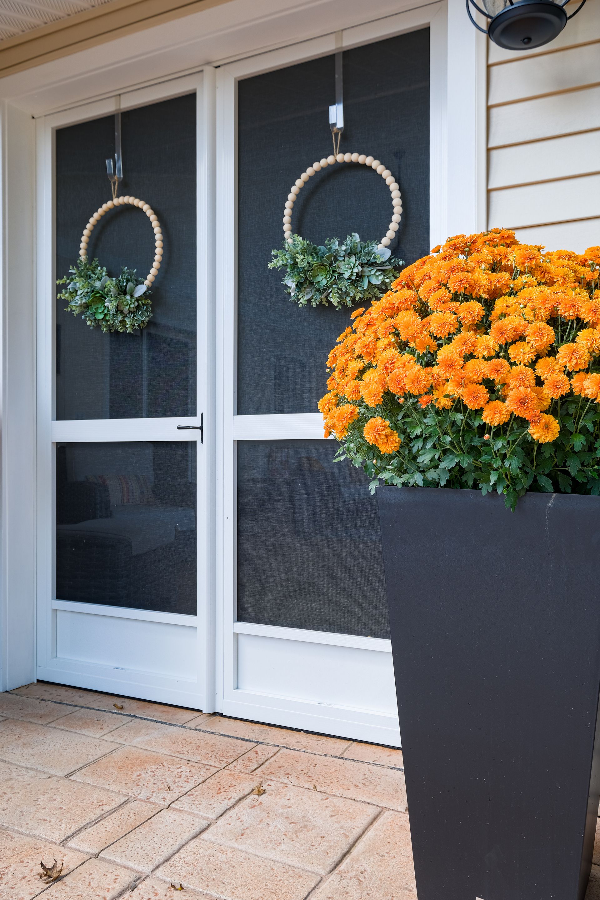 Two screen doors decorated with wreaths. Orange mums in a tall black pot.
