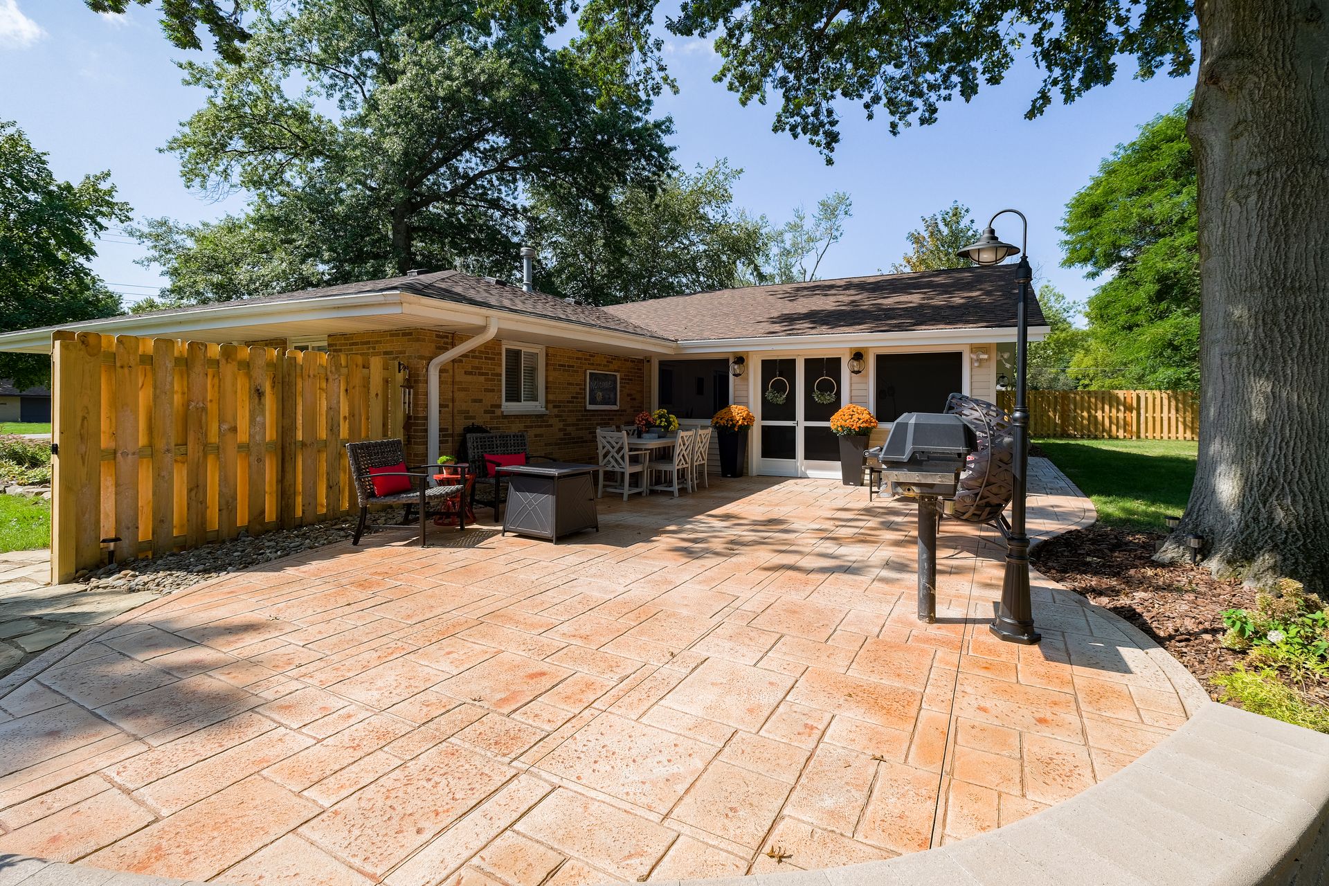 Patio with brick-like paving, covered seating area, grill, and a house. Sunny day.