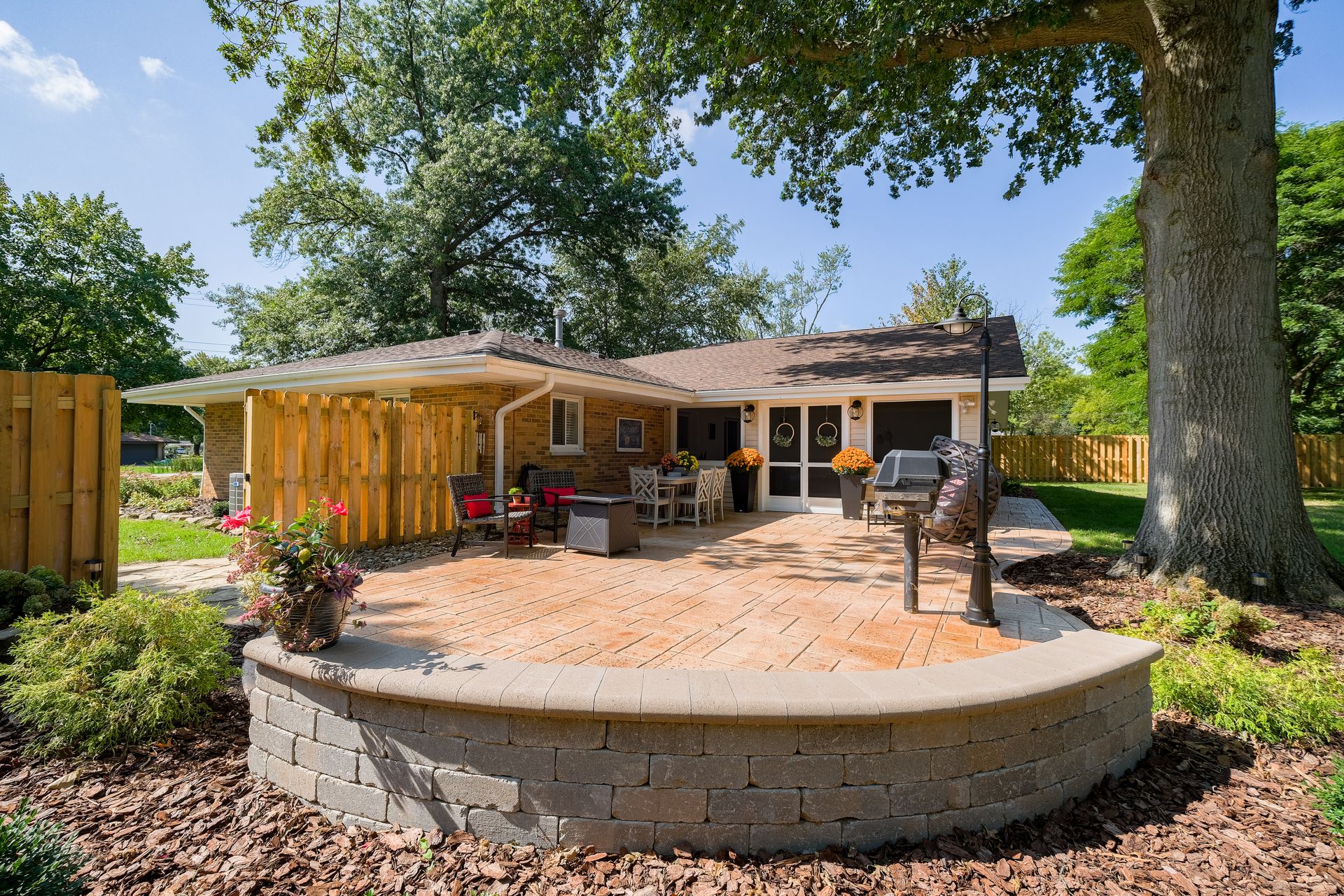 Backyard patio with brick retaining wall, grill, and seating area under a wooden canopy.