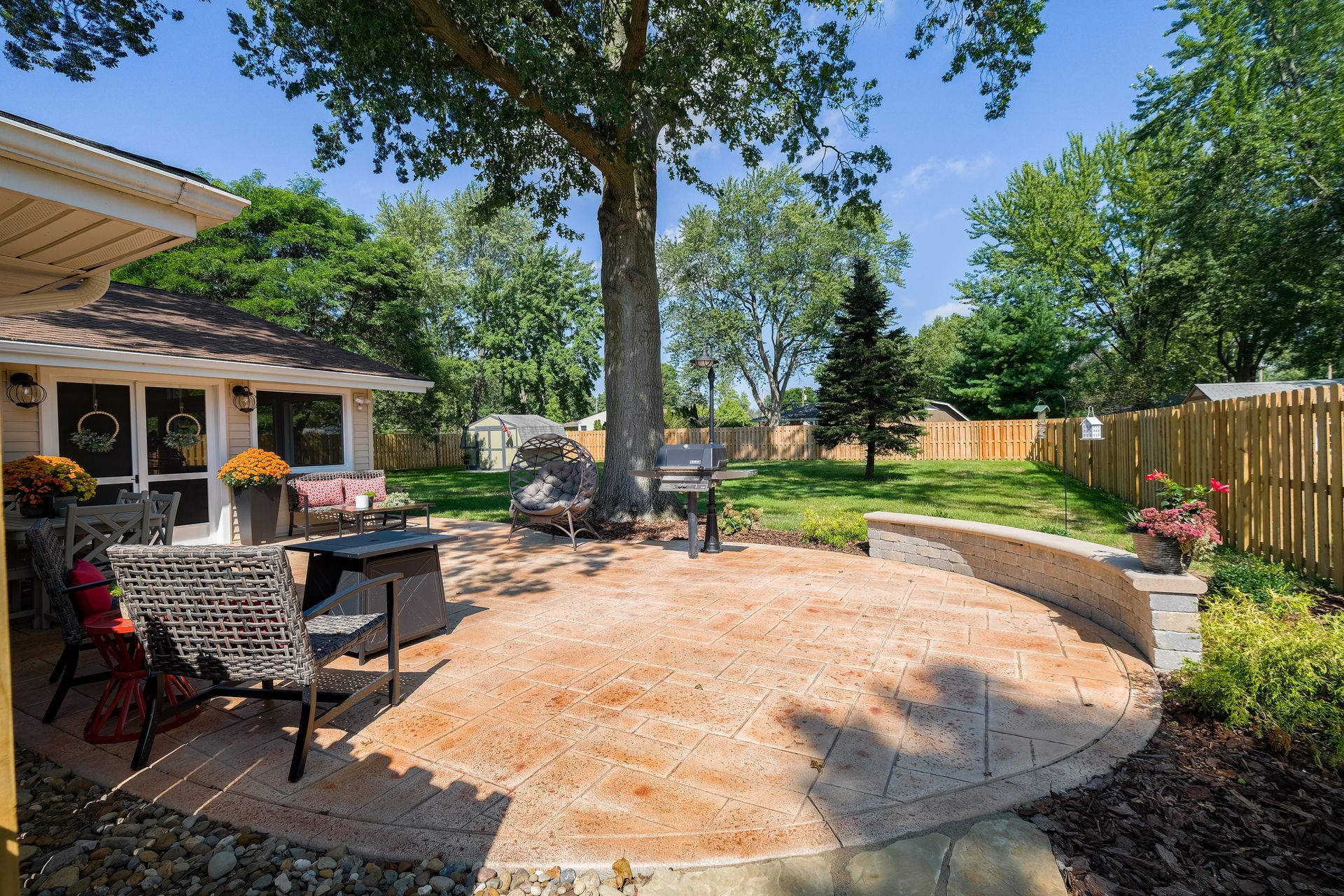 Backyard patio with seating, grill, and a curved retaining wall. Sunny day with trees and a wooden fence.