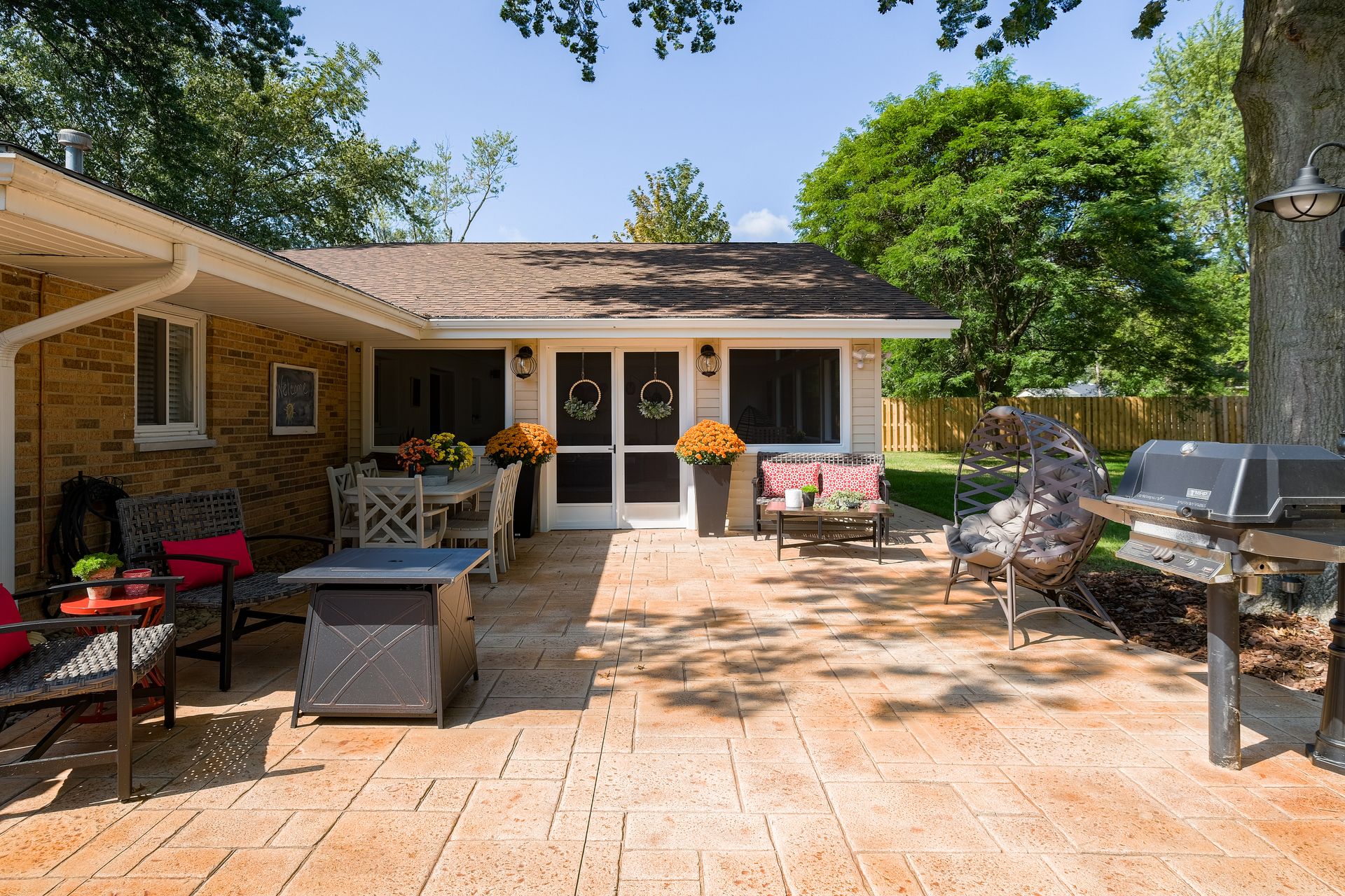 Patio with brick building, dining table, seating, grill, and lush trees in the background.