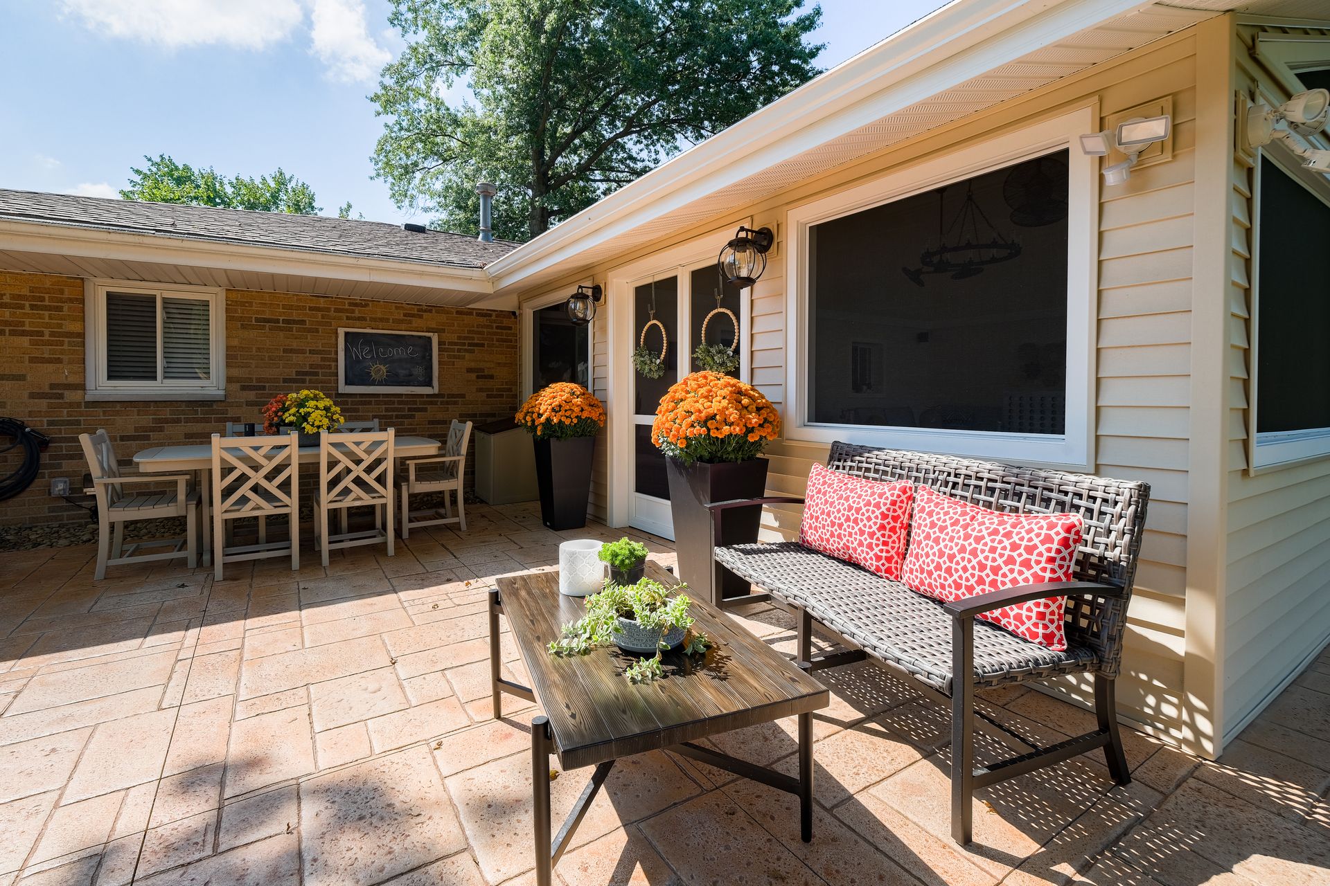 Patio with table, chairs, bench, and potted flowers. Beige brick walls, tan tiled floor. Bright sunny day.