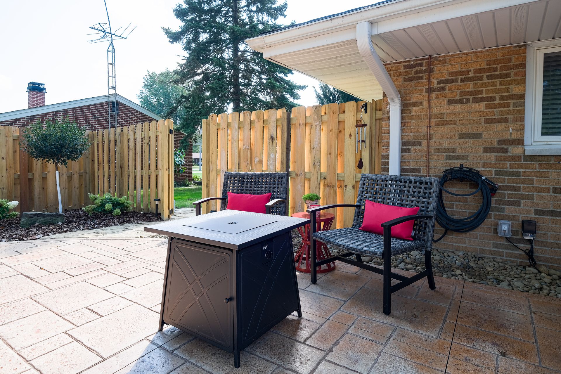 Patio with two chairs, fire pit, and wooden fence. Red pillows pop against patterned seats. Brick house exterior.