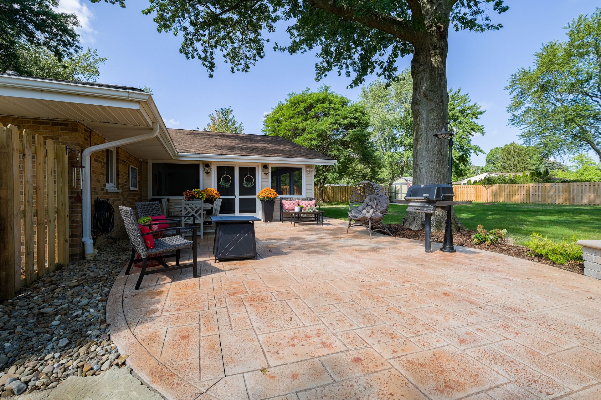 Patio with stamped concrete, grill, chairs, and small building under a tree in a backyard.