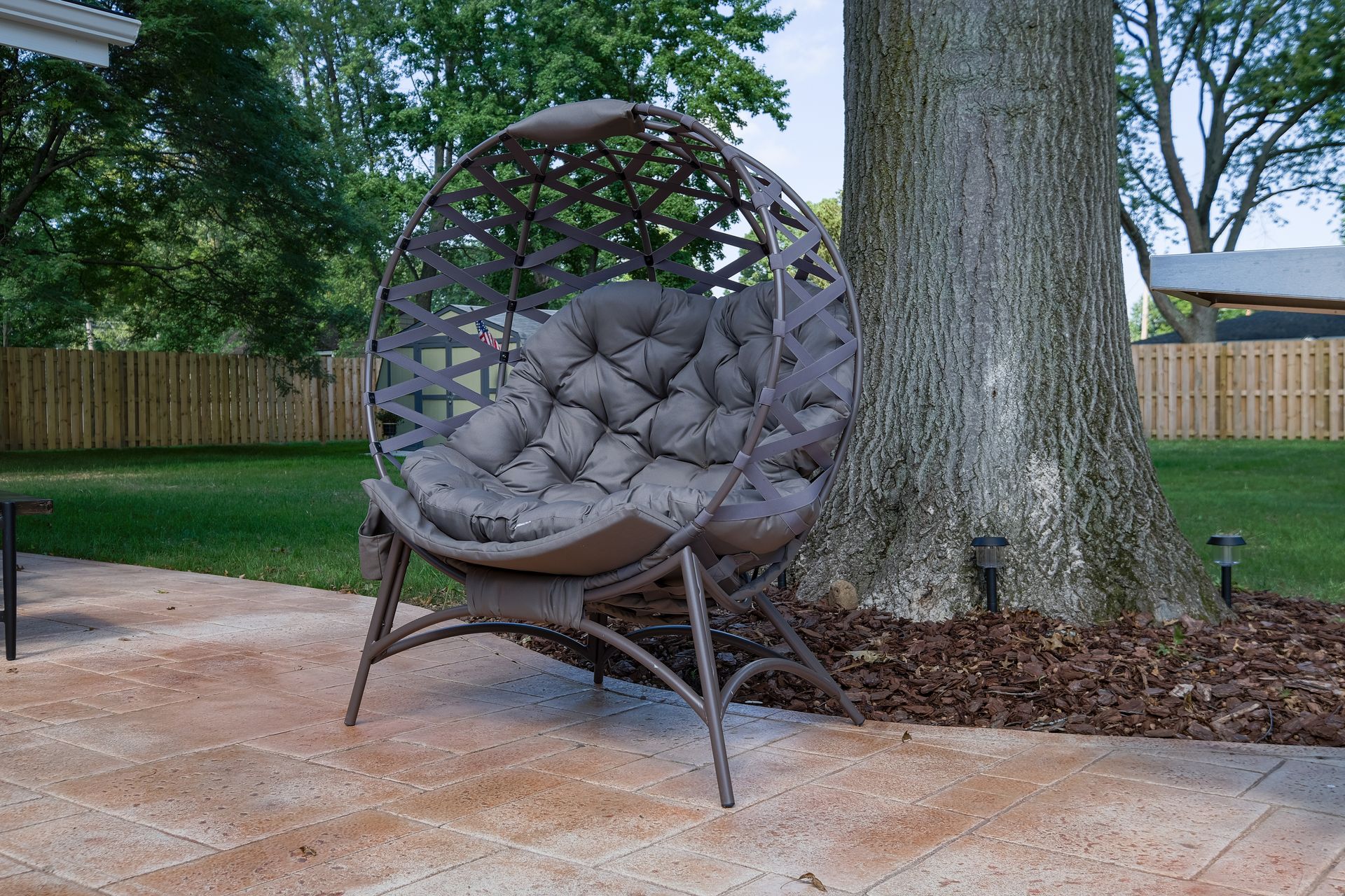 Wicker egg chair with gray cushions sits on a brick patio next to a tree. Green grass and wooden fence in the background.