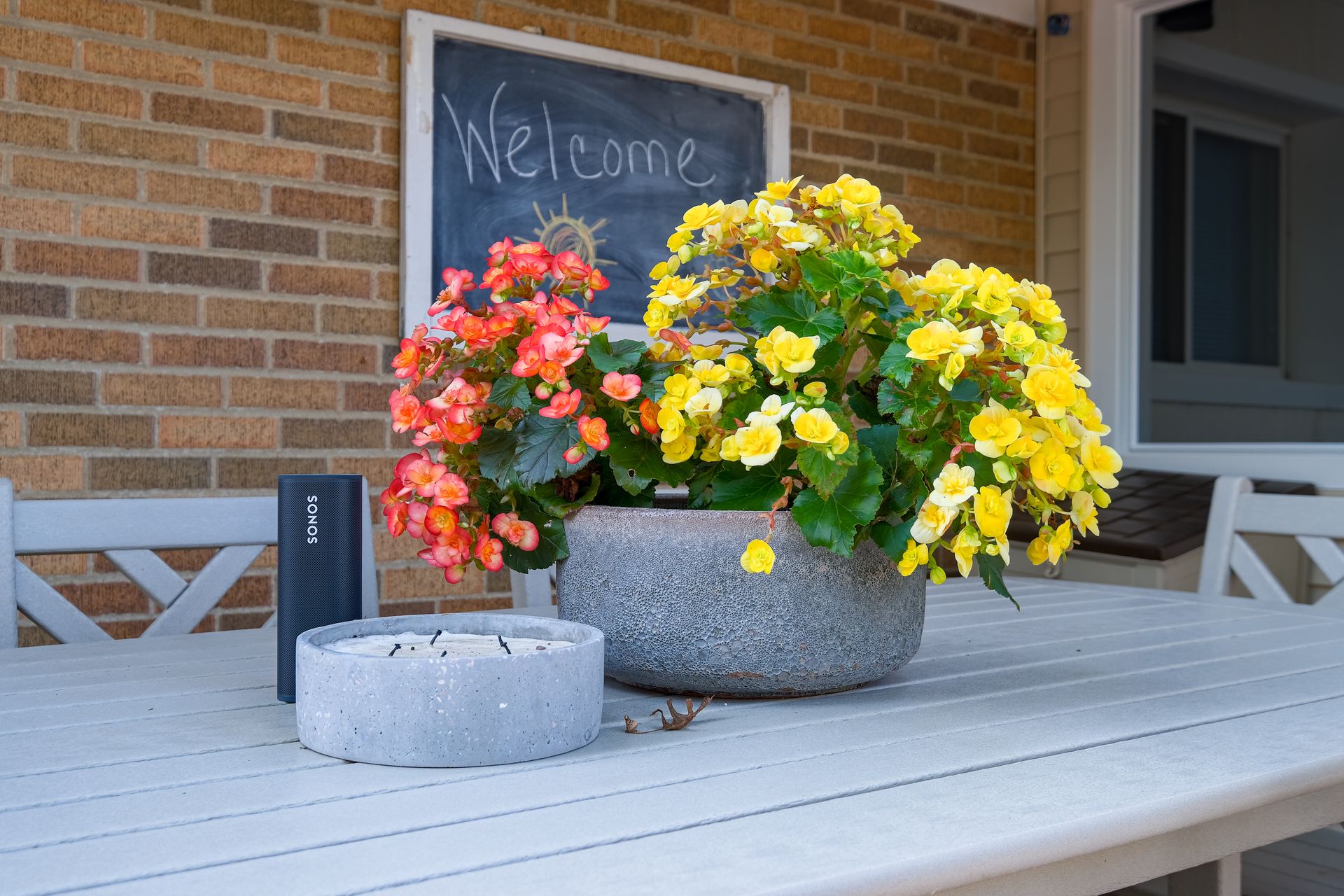 Flowers in a stone planter on a white table. An Amazon Echo, and candle sit on the table. Welcome sign on brick wall.