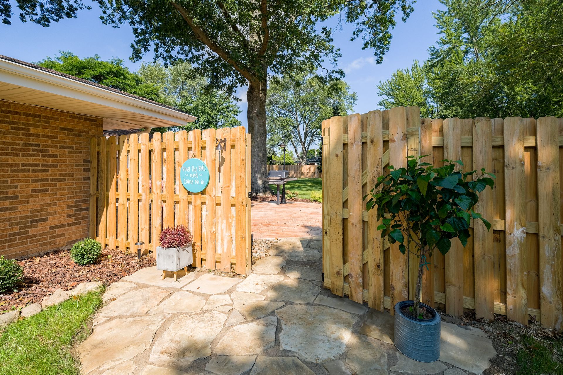 Wooden fence gate leading to a brick patio and garden area with plants and trees.