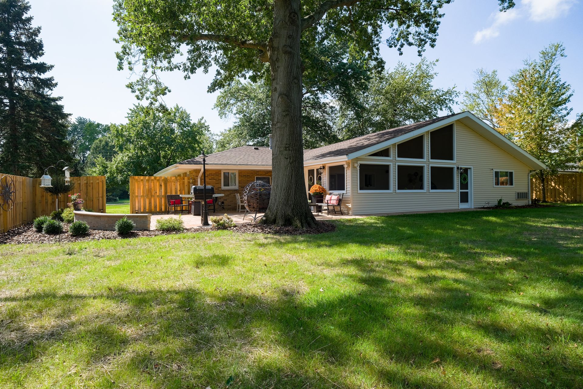 Backyard with house, wooden fence, grill, tree, and green grass on a sunny day.