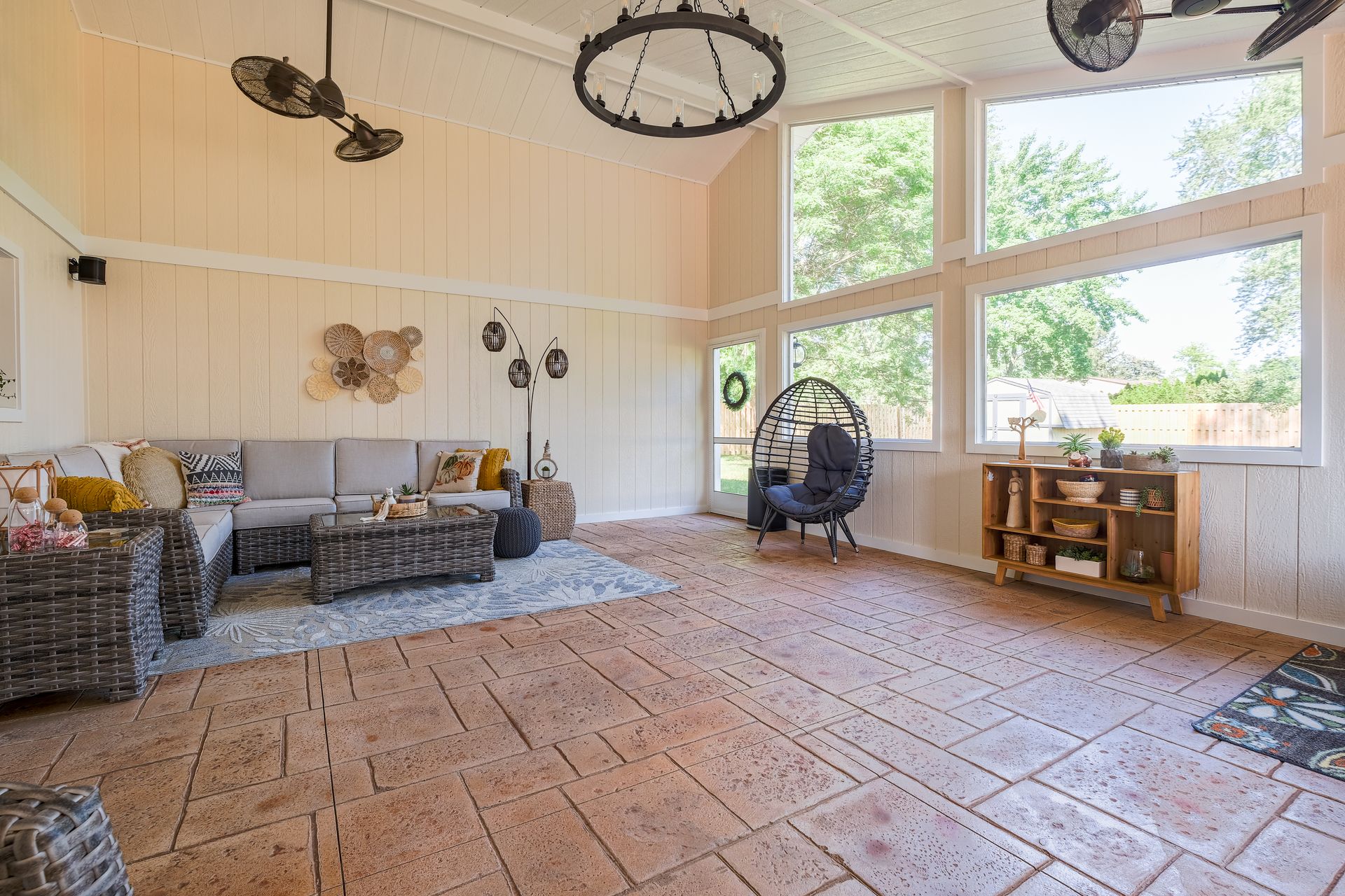 Cozy sunroom with gray sofa, large windows, and brick-like flooring, overlooking a green yard.
