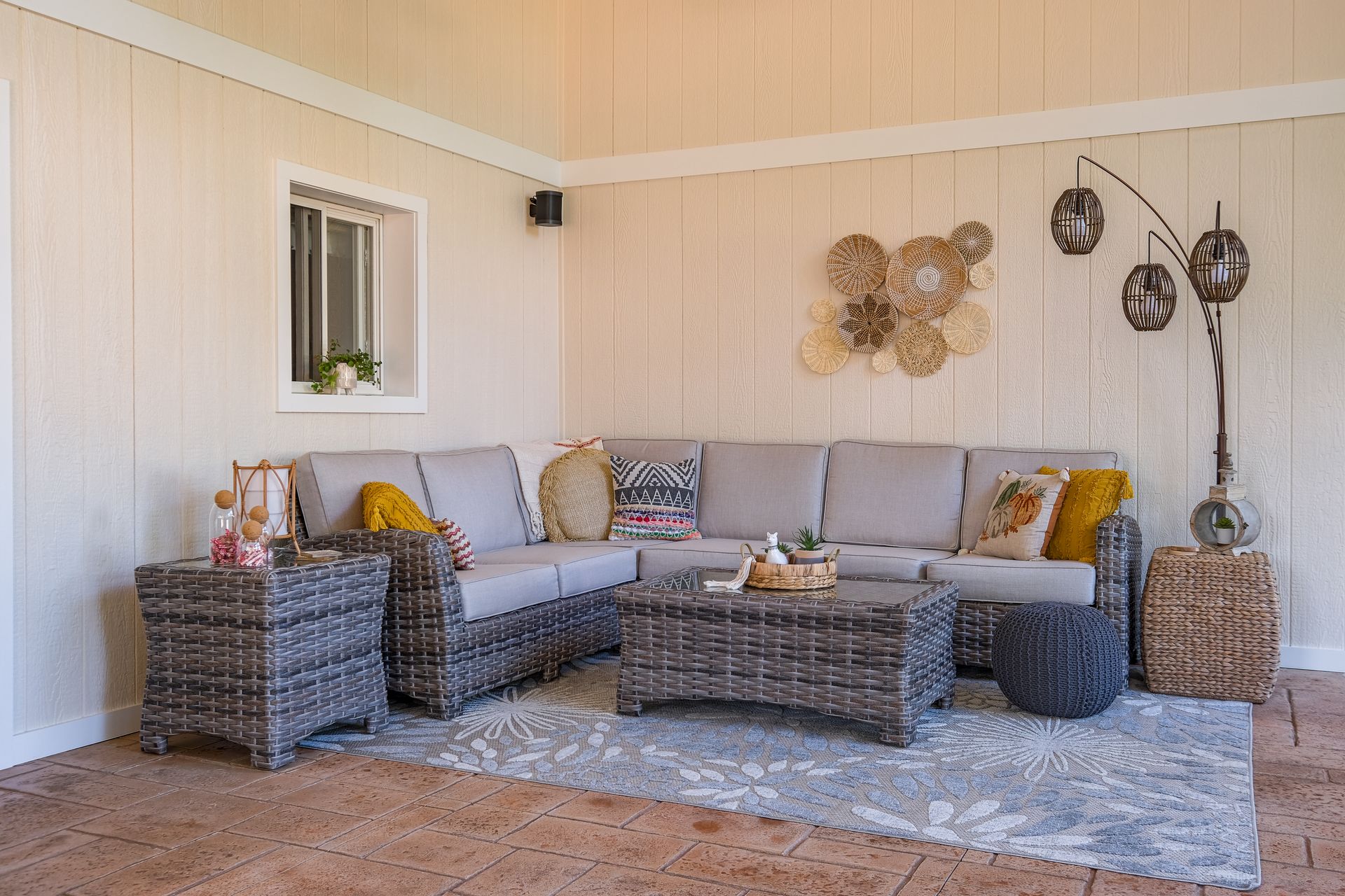 L-shaped gray wicker outdoor seating on a rug in a covered patio with decorative wall art and plants.