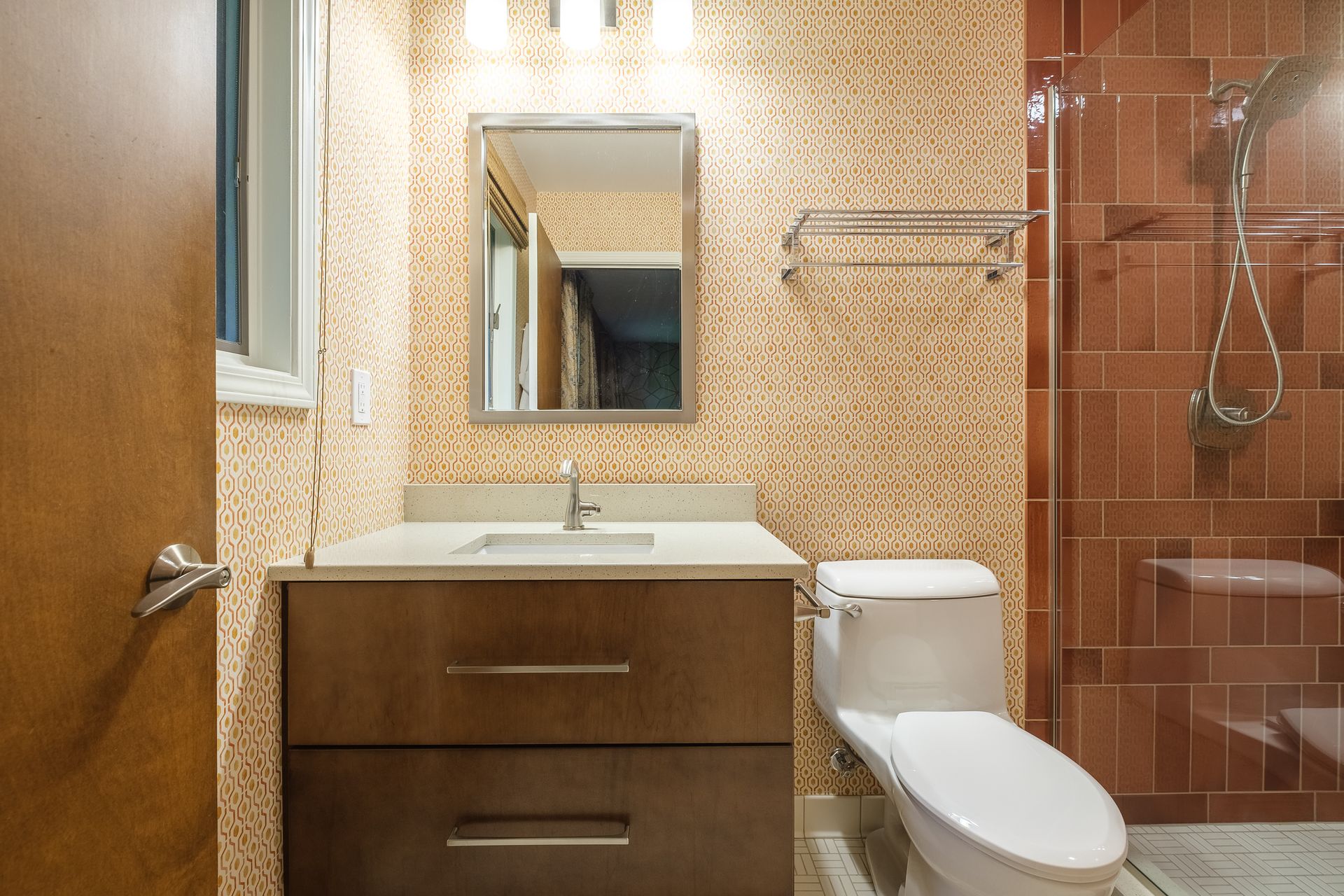 Bathroom with wood vanity, patterned wallpaper, toilet, and a tiled shower.
