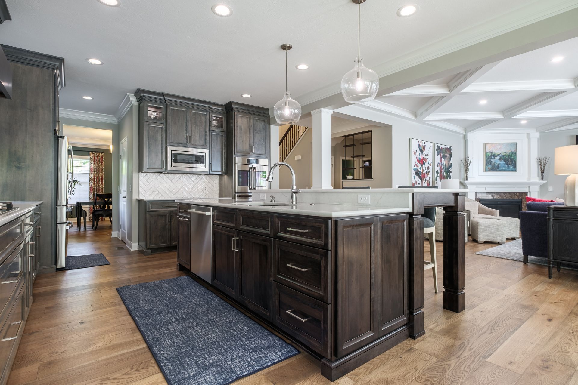 Dark-wood kitchen with island, stainless steel appliances, and white countertops. View into the living area with fireplace.