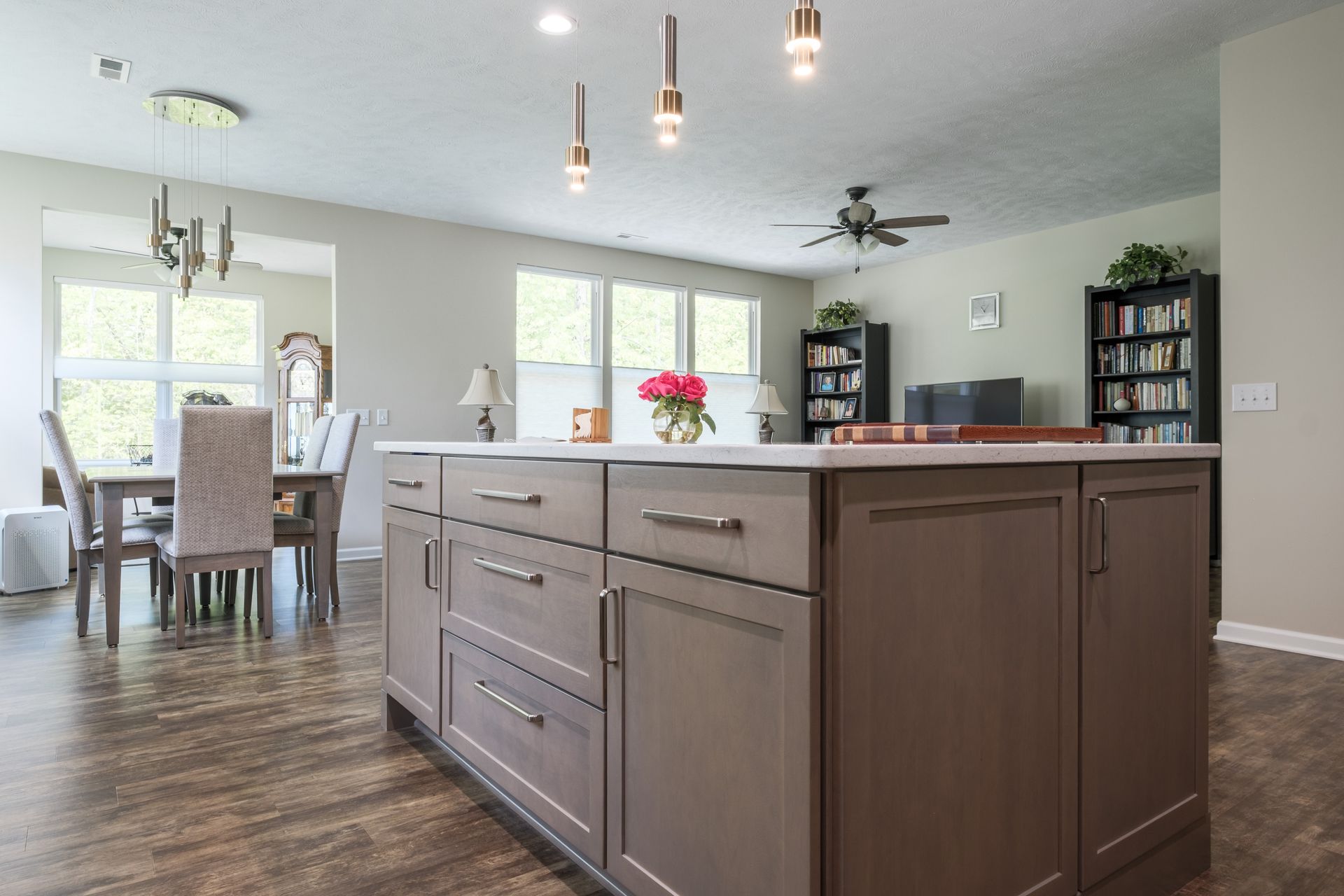 Kitchen with island, dining area, and living room. Brown cabinets, wooden floors, windows, and light fixtures.