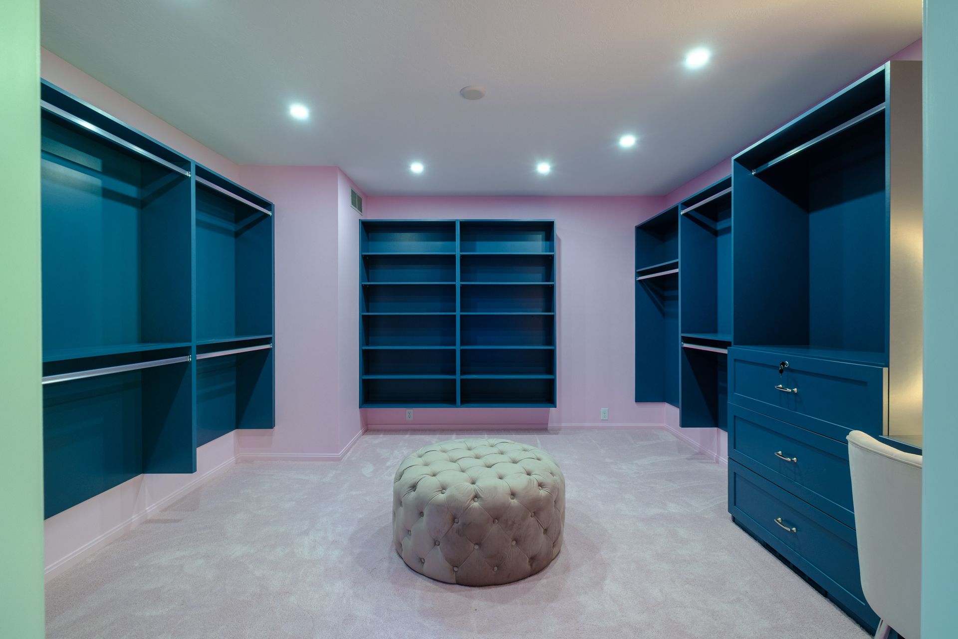 Pink and teal walk-in closet with shelves, drawers, and an ottoman on a pink carpet.