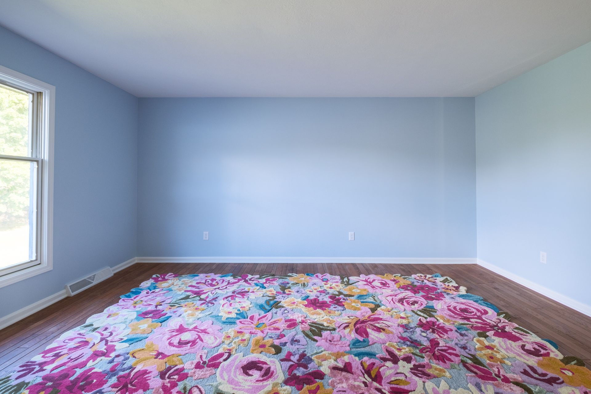 Empty room with blue walls, wood floor, floral rug, and window.