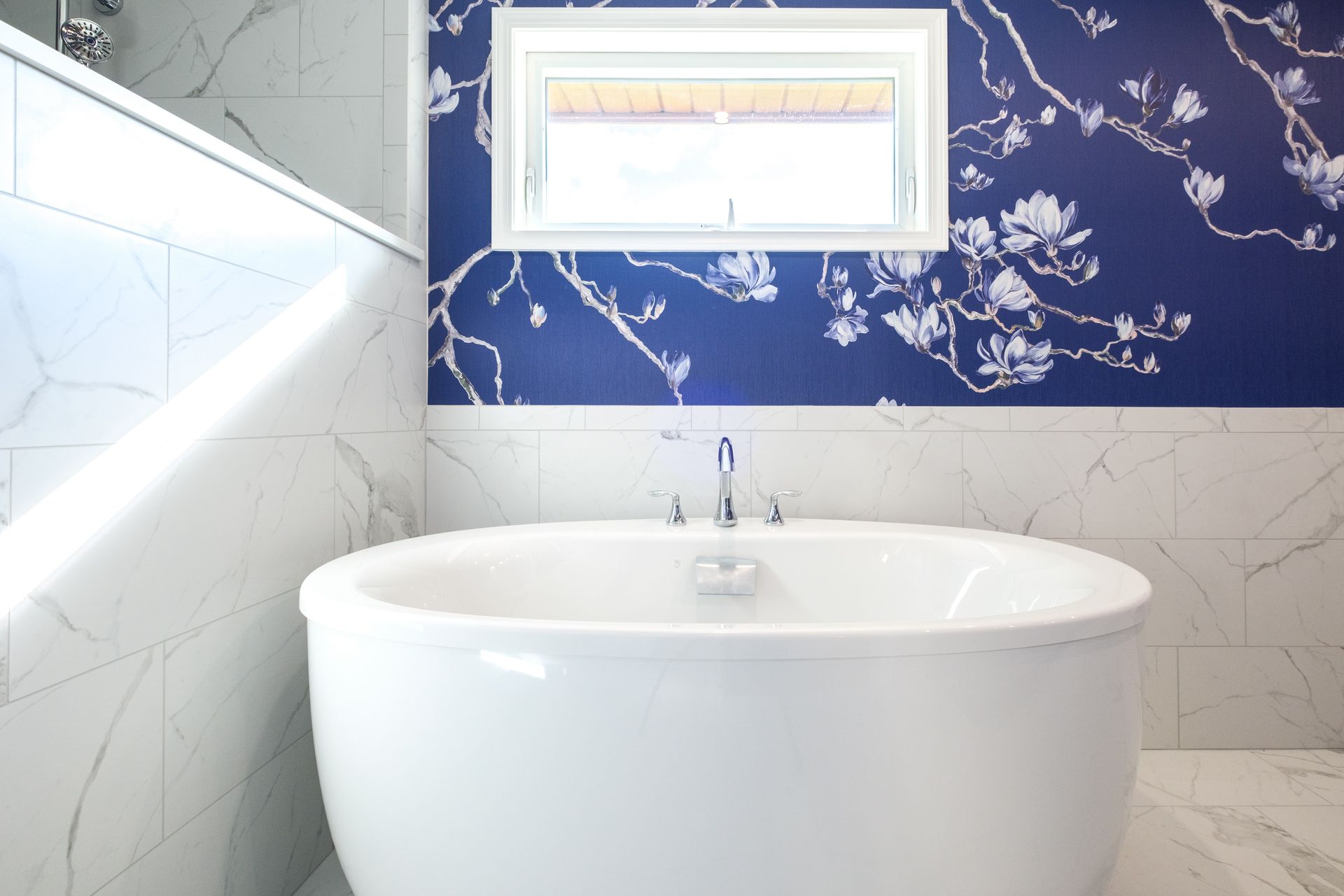 Oval bathtub in a bathroom with a blue floral wallpaper accent wall and a window.