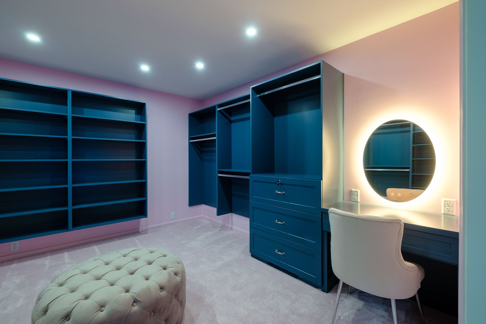 Blue closet with shelves, drawers, and hanging rods; a vanity, and a tufted ottoman in a pink room.