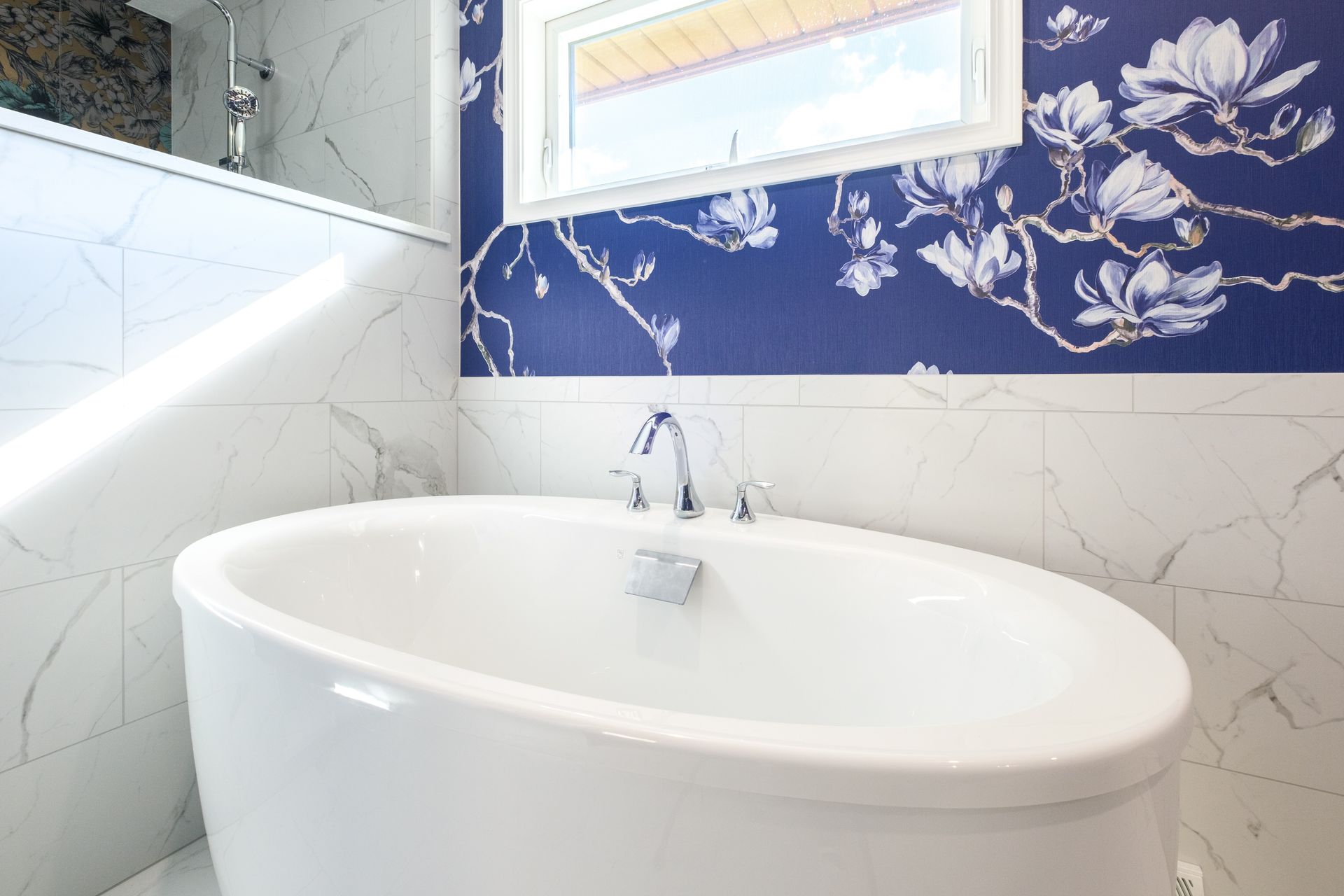 White oval bathtub in a bathroom with blue floral wallpaper and a small window.