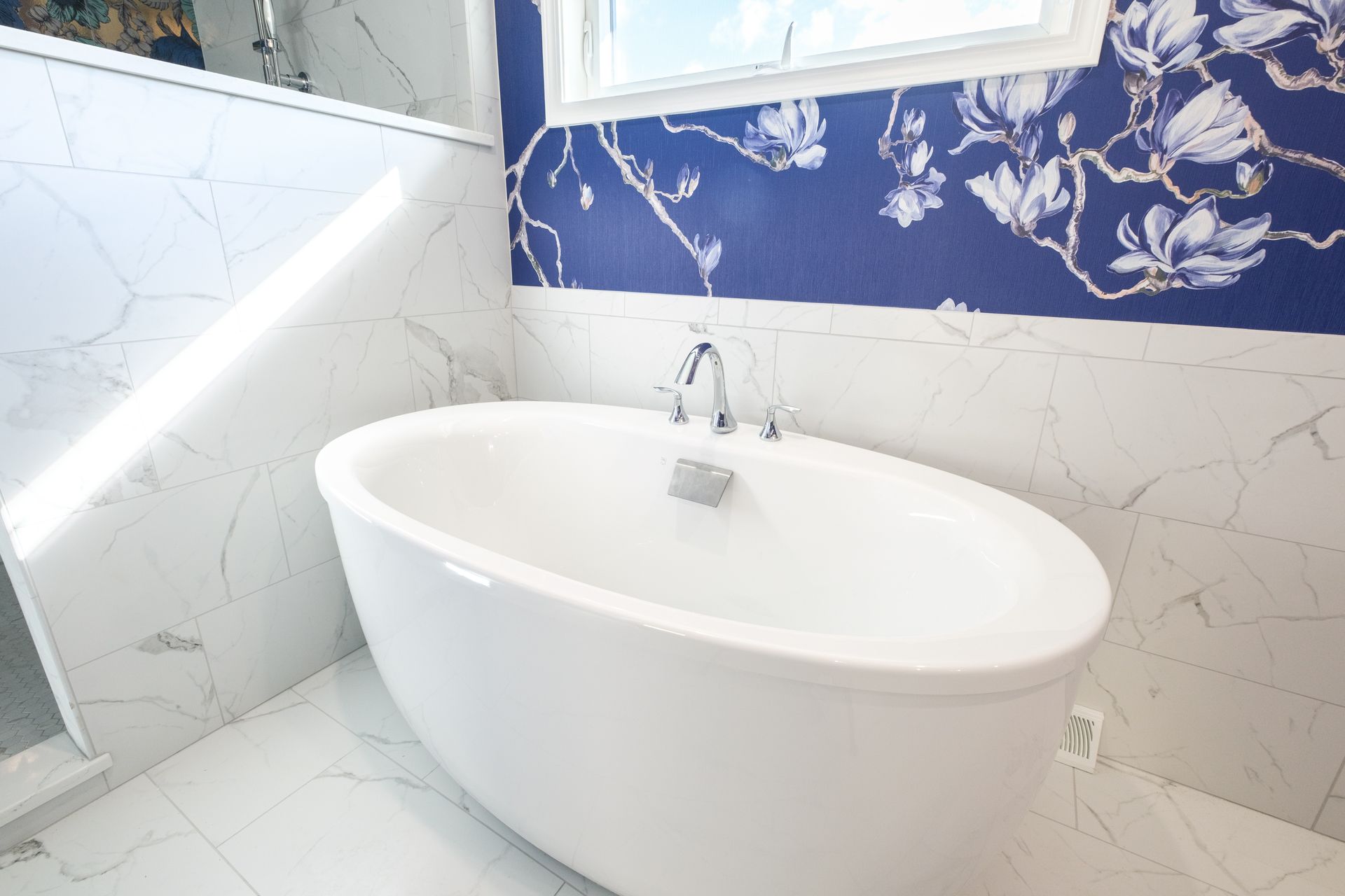 White oval bathtub in a bathroom with marble tile and blue floral wallpaper.