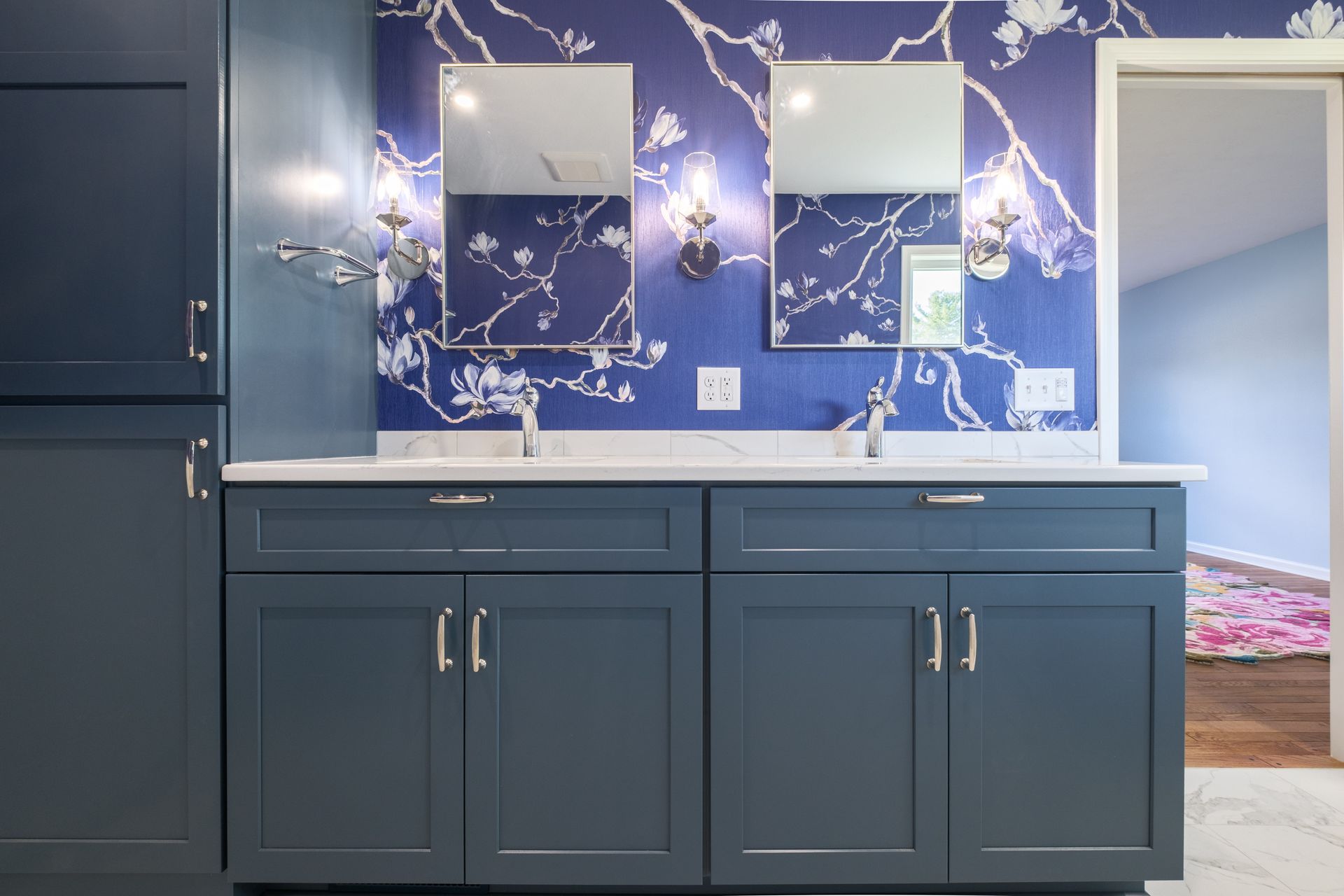 Blue bathroom with double vanity, floral wallpaper, and tall storage cabinet.