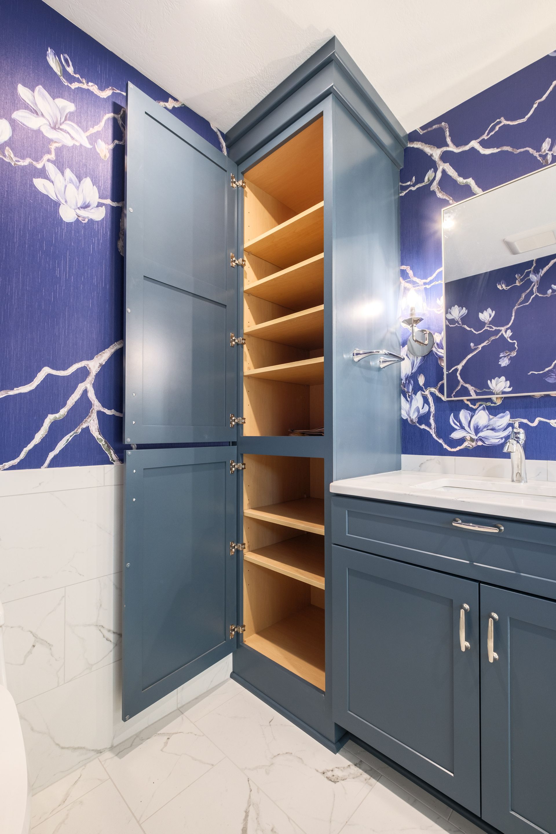 Blue bathroom storage cabinet with open doors, wooden shelves. Blue floral wallpaper.