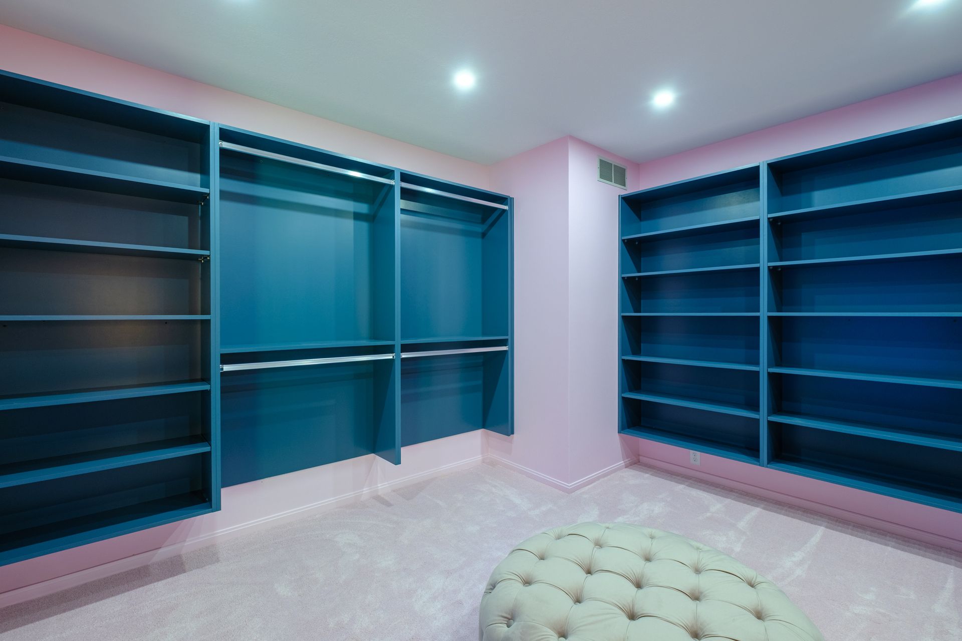 A walk-in closet with teal shelves and pink walls, featuring an upholstered ottoman.