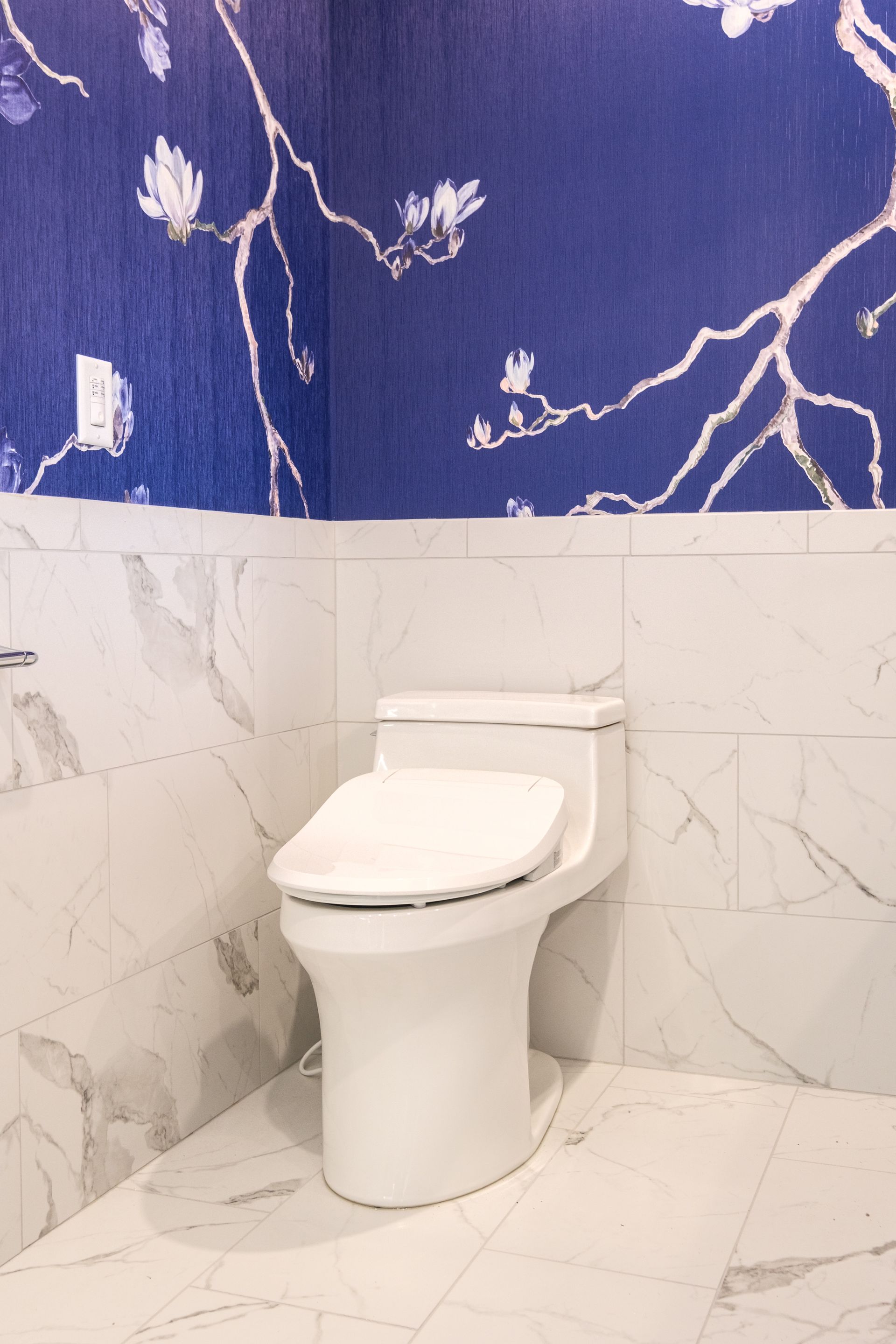 White toilet in a bathroom with blue floral wallpaper and white marble tile.